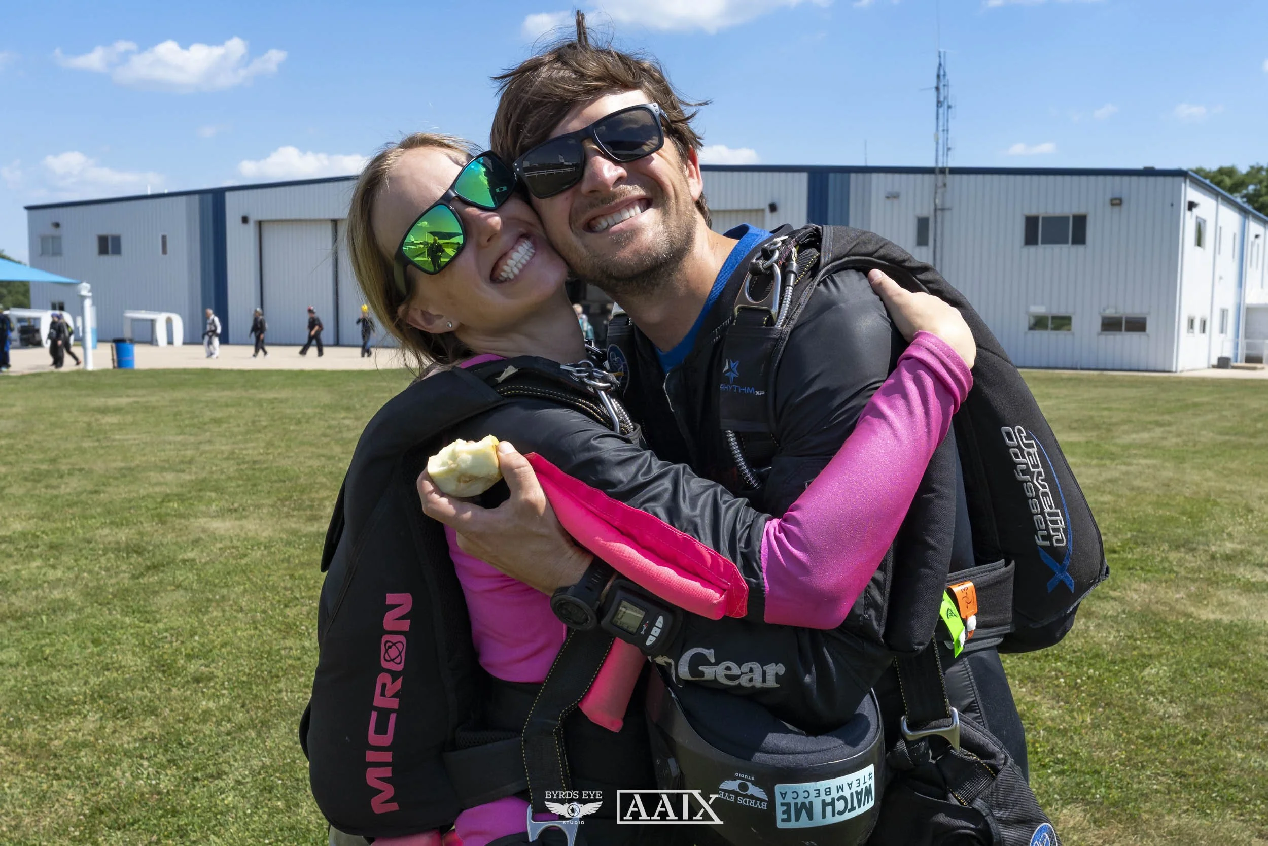 Two skydivers in jumpsuits hugging and smiling at a skydiving center on a sunny day, with a large white hangar building in the background.