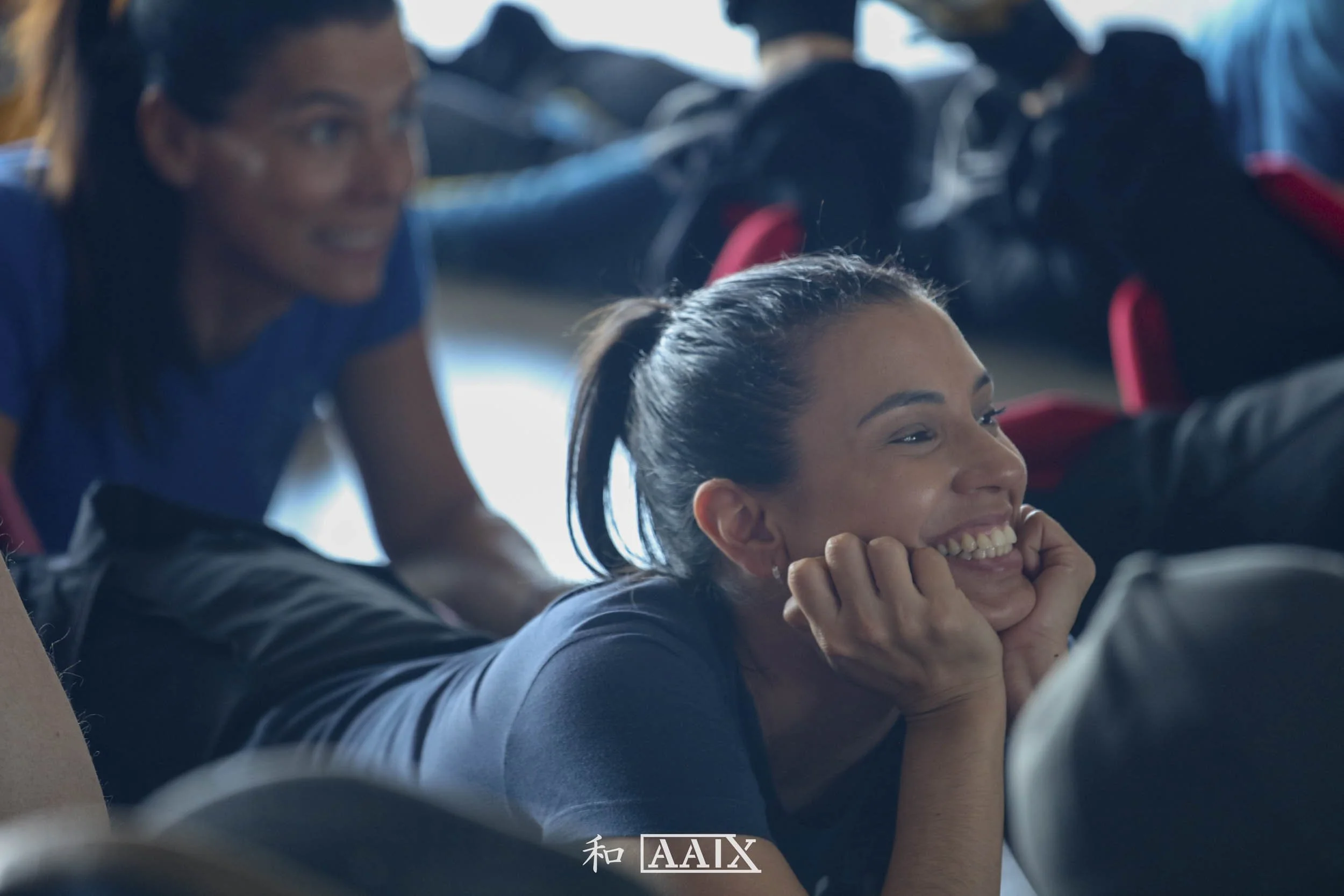 Two women in a casual setting, one lying down and smiling with her chin resting on her hands, the other leaning on her hands and smiling, with a blurry background of people and bags.