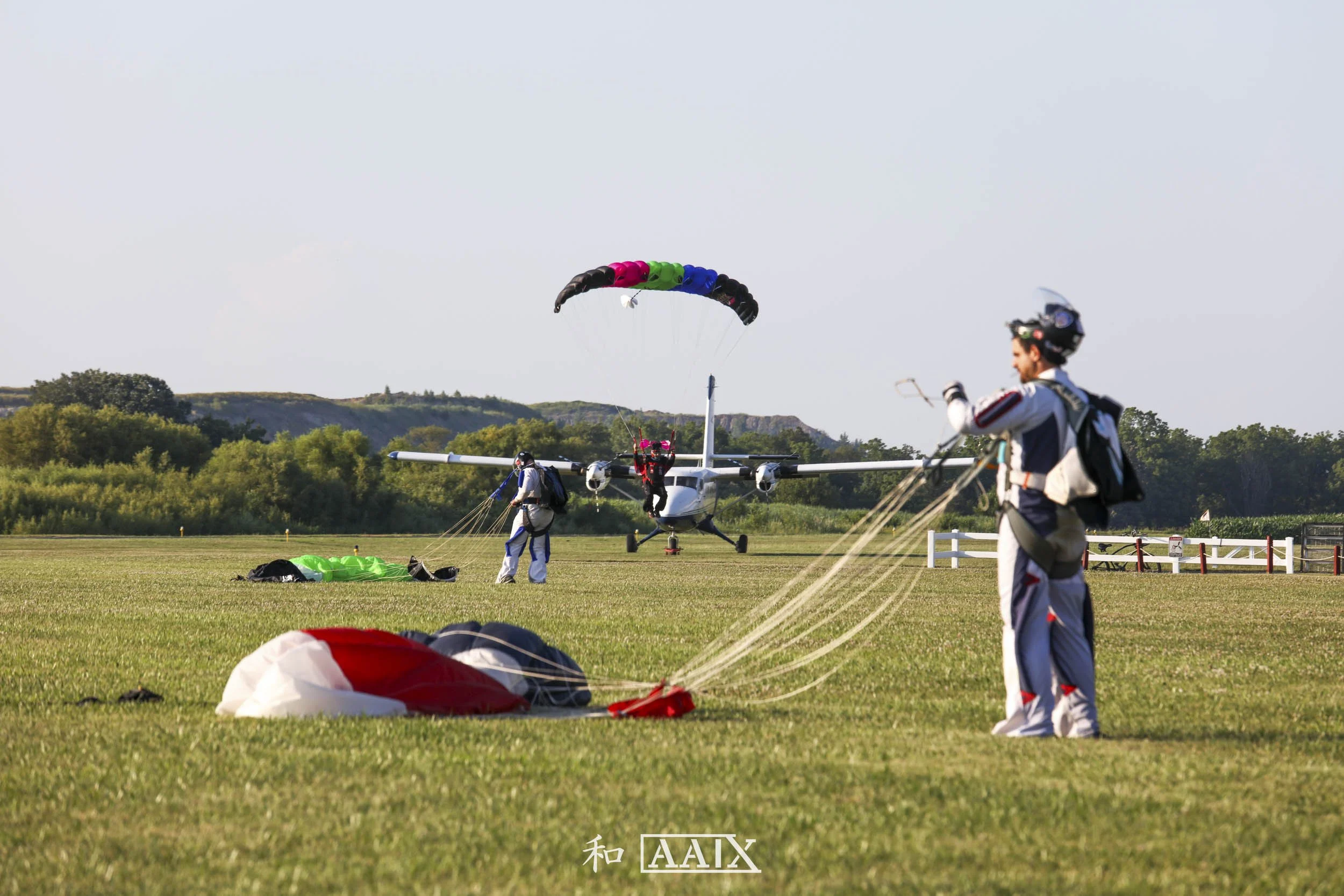 Skydivers preparing for departure on an airfield with a small airplane in the background, some skydivers are on the ground handling parachutes while one is mid-air with a colorful parachute.