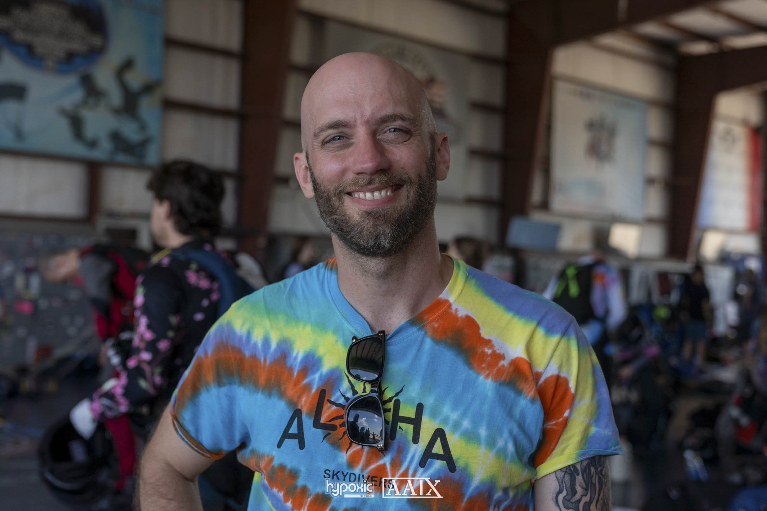 A smiling man with a beard and bald head wearing a colorful tie-dye t-shirt with the word 'Aloha' and sunglasses hanging from the neckline, inside a busy indoor space.