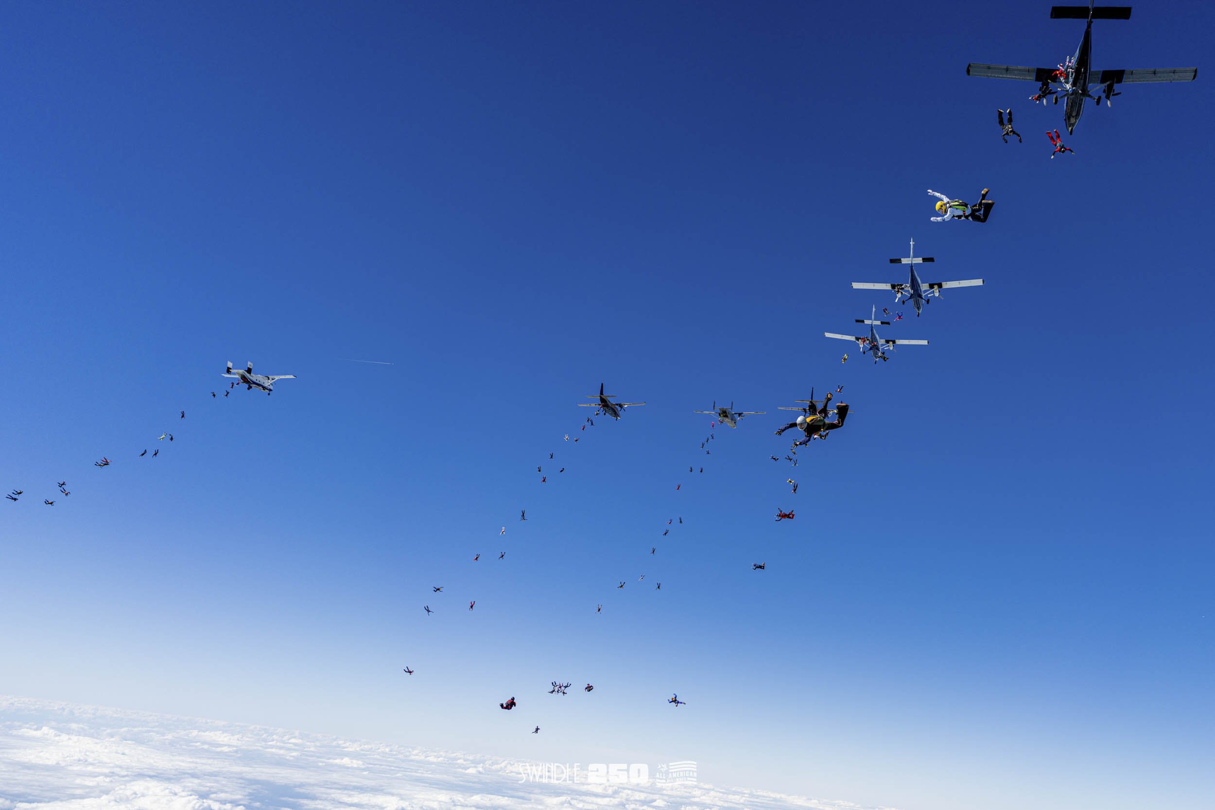 Group of skydivers jumping from helicopters, free-falling against a clear blue sky over a snowy landscape.