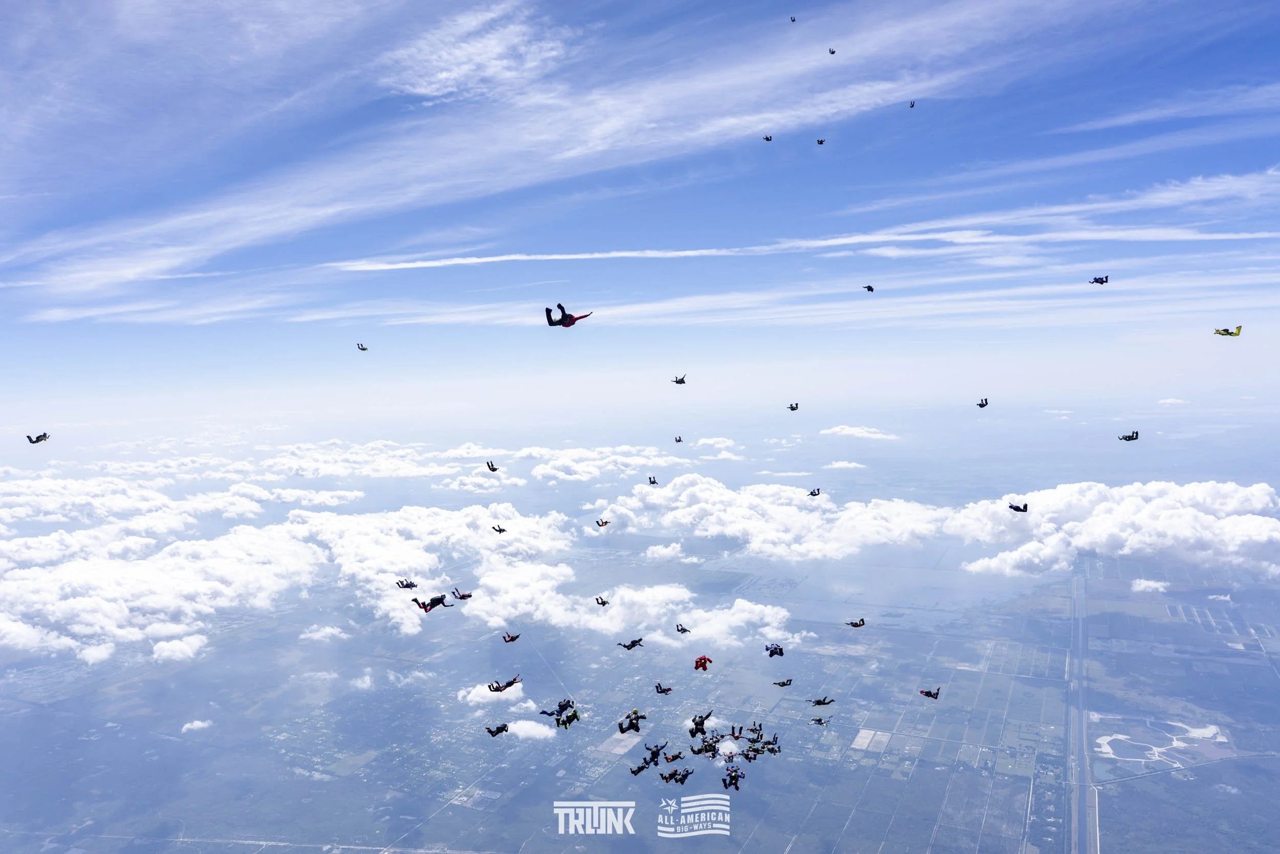 Multiple skydivers falling through the sky over clouds and the ground, with some skydivers in bright suits and a blue sky with wispy clouds.