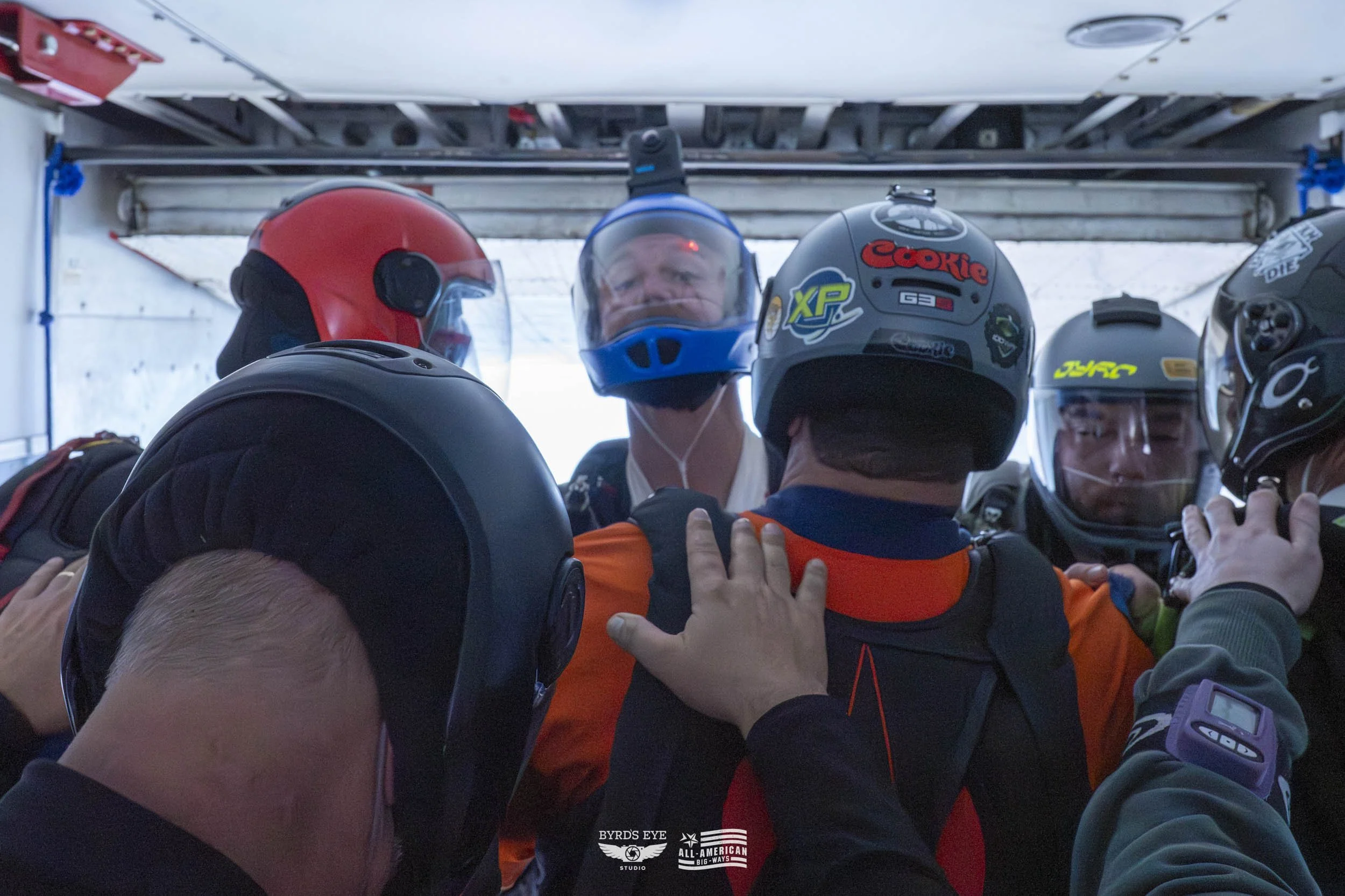 Group of people wearing motorcycle helmets and gear, standing close together inside a vehicle or trailer.