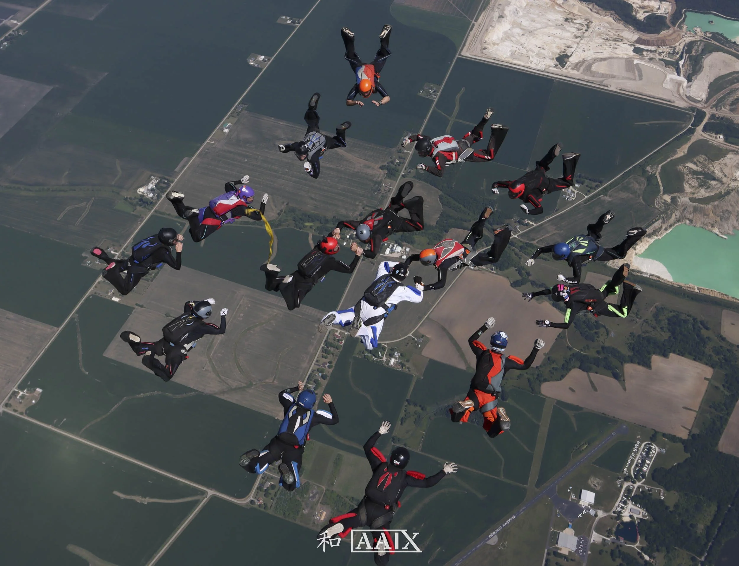 A group of skydivers in jumpsuits and helmets free-falling over a rural landscape with fields, roads, and water bodies.