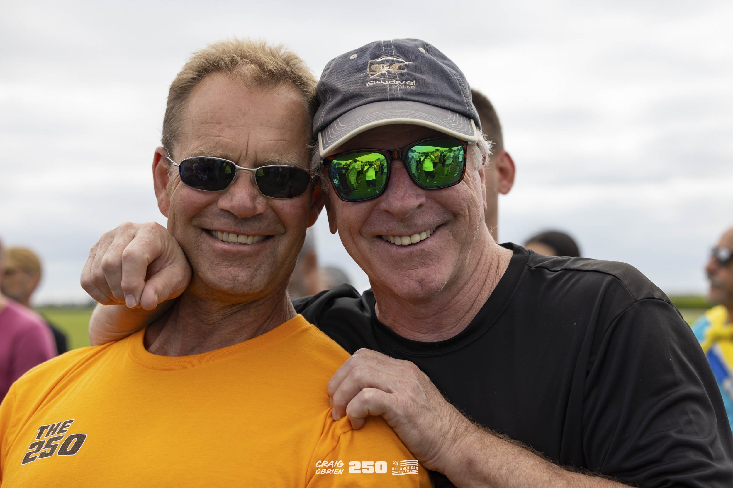 Two smiling men wearing sunglasses, one in a yellow shirt with 'The 250' logo and the other in a black shirt and cap, pose outdoors on a cloudy day.