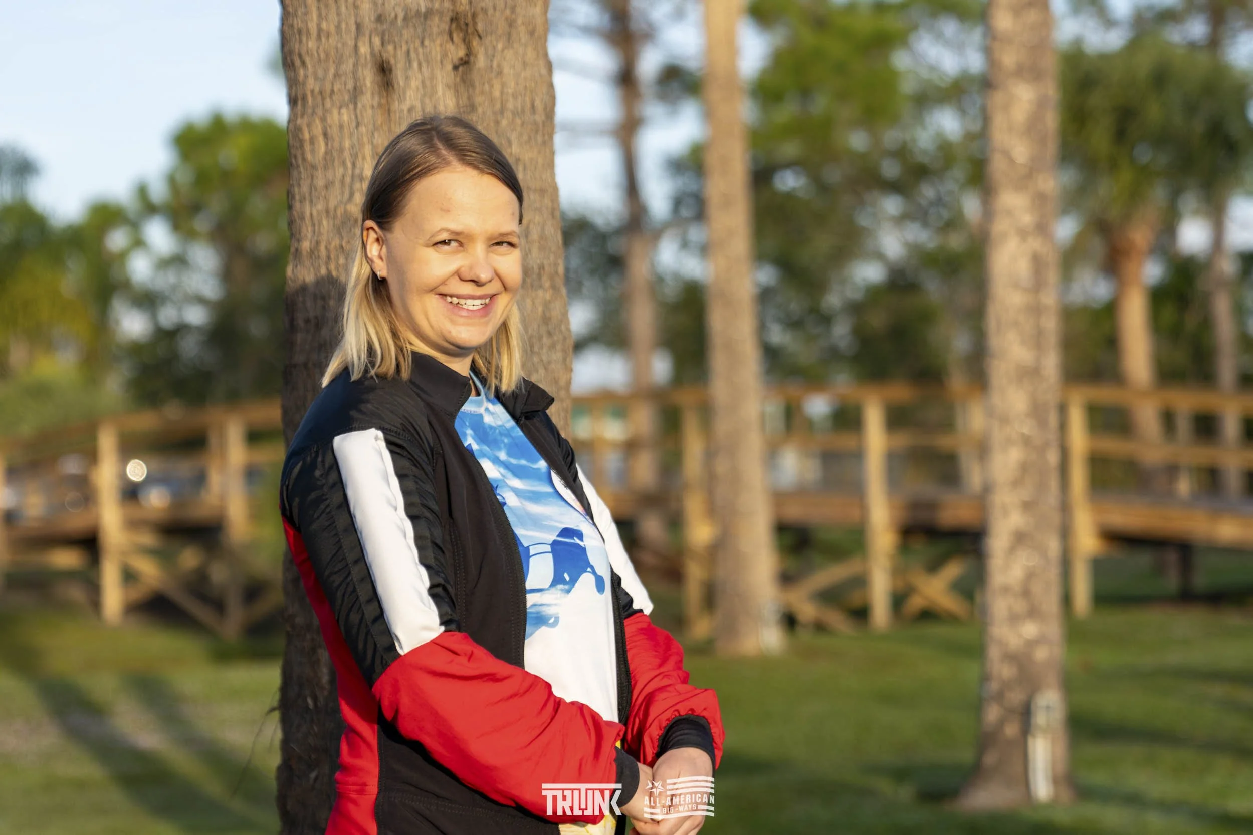 A woman with shoulder-length blonde hair smiling outdoors, leaning against a tree, wearing a black, red, white, and blue jacket with a graphic t-shirt featuring a scenic design, with a wooden walkway and trees in the background.