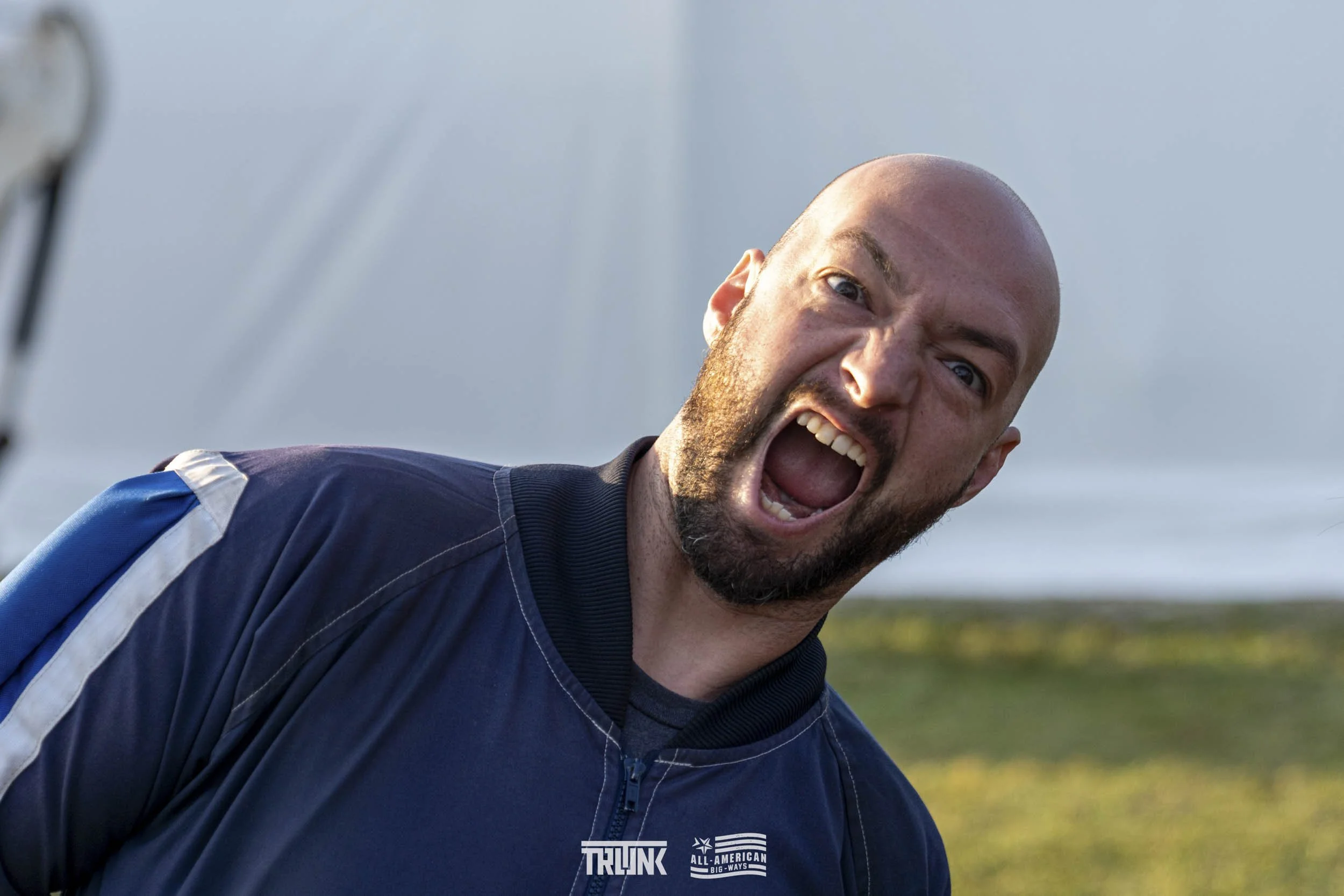 A man with a bald head and beard wearing a navy blue jacket with logos, shouting or yelling outdoors.