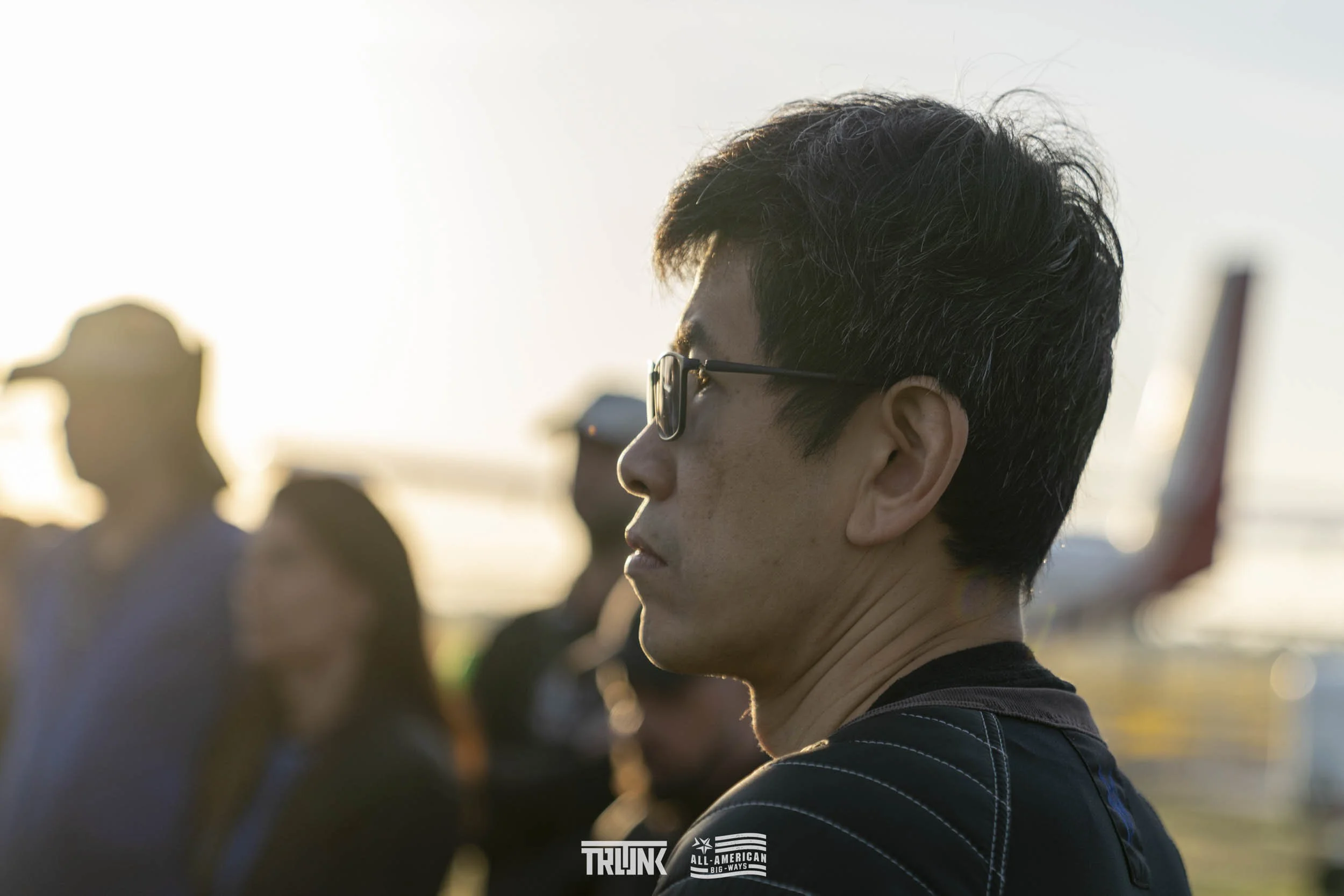 A man with black hair and glasses standing outdoors during sunset, surrounded by blurred people and an airplane in the background.