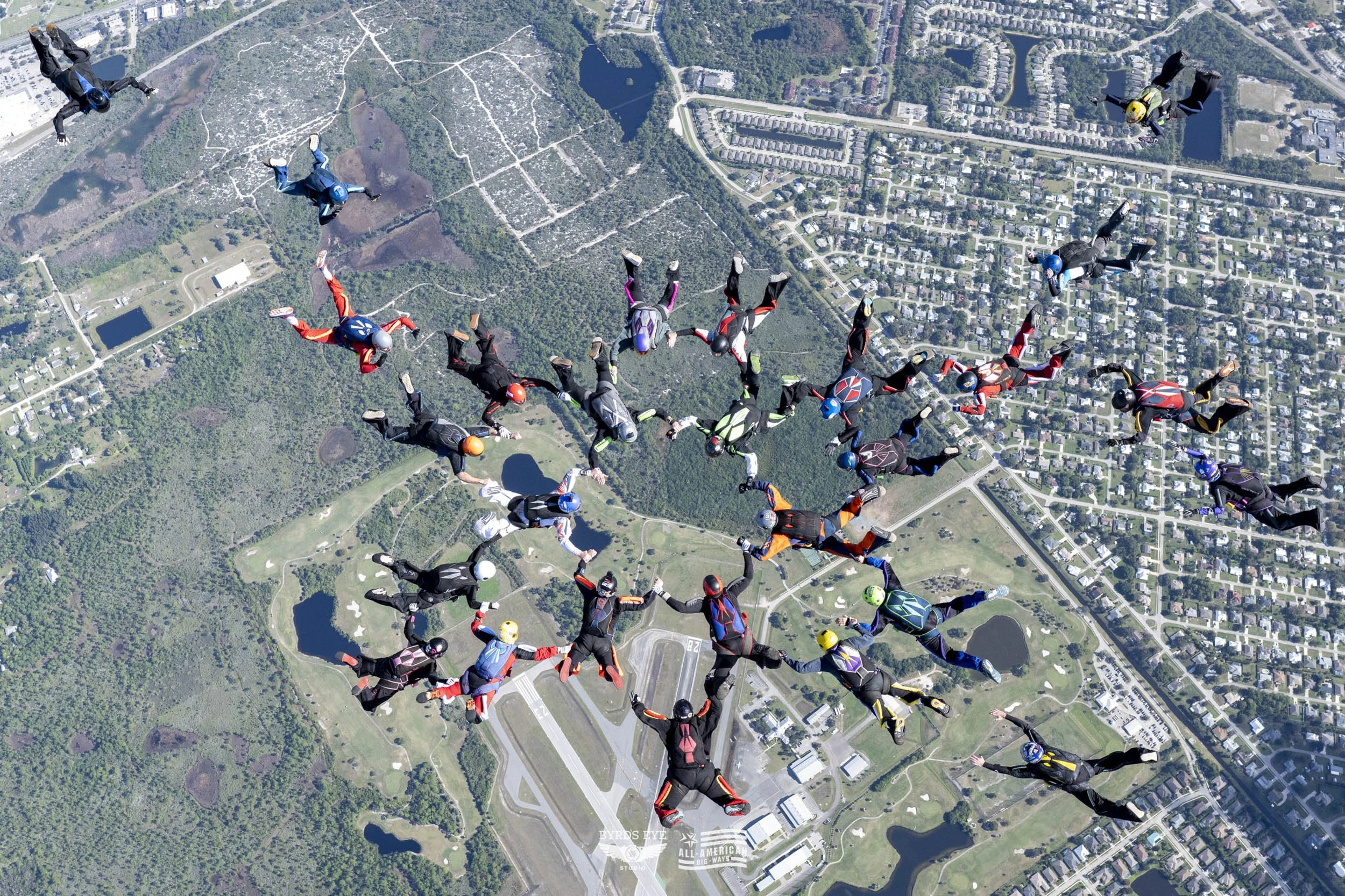 Group of skydivers in free fall above a landscape with lakes, forests, and a residential area.