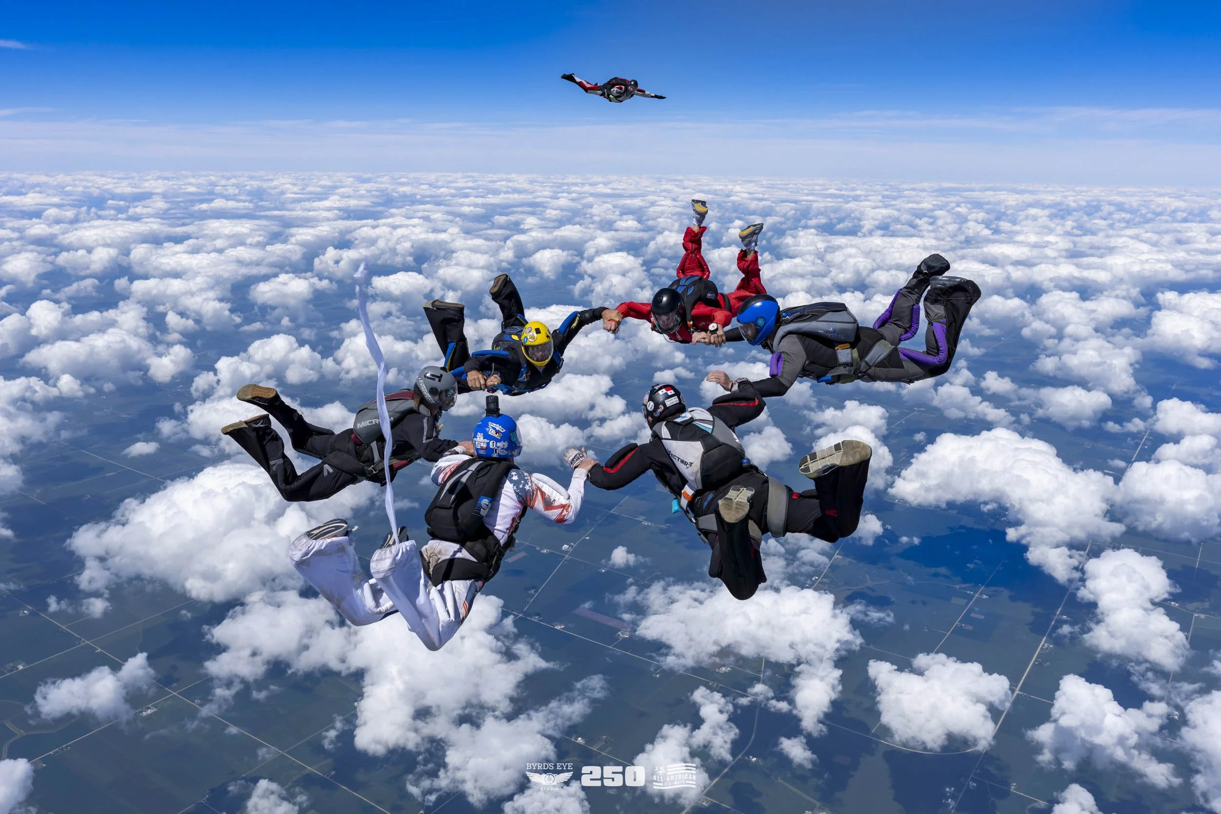 Eight skydivers in freefall holding hands in a circle above the clouds during daytime, with one skydiver in a head-down position closer to the camera.