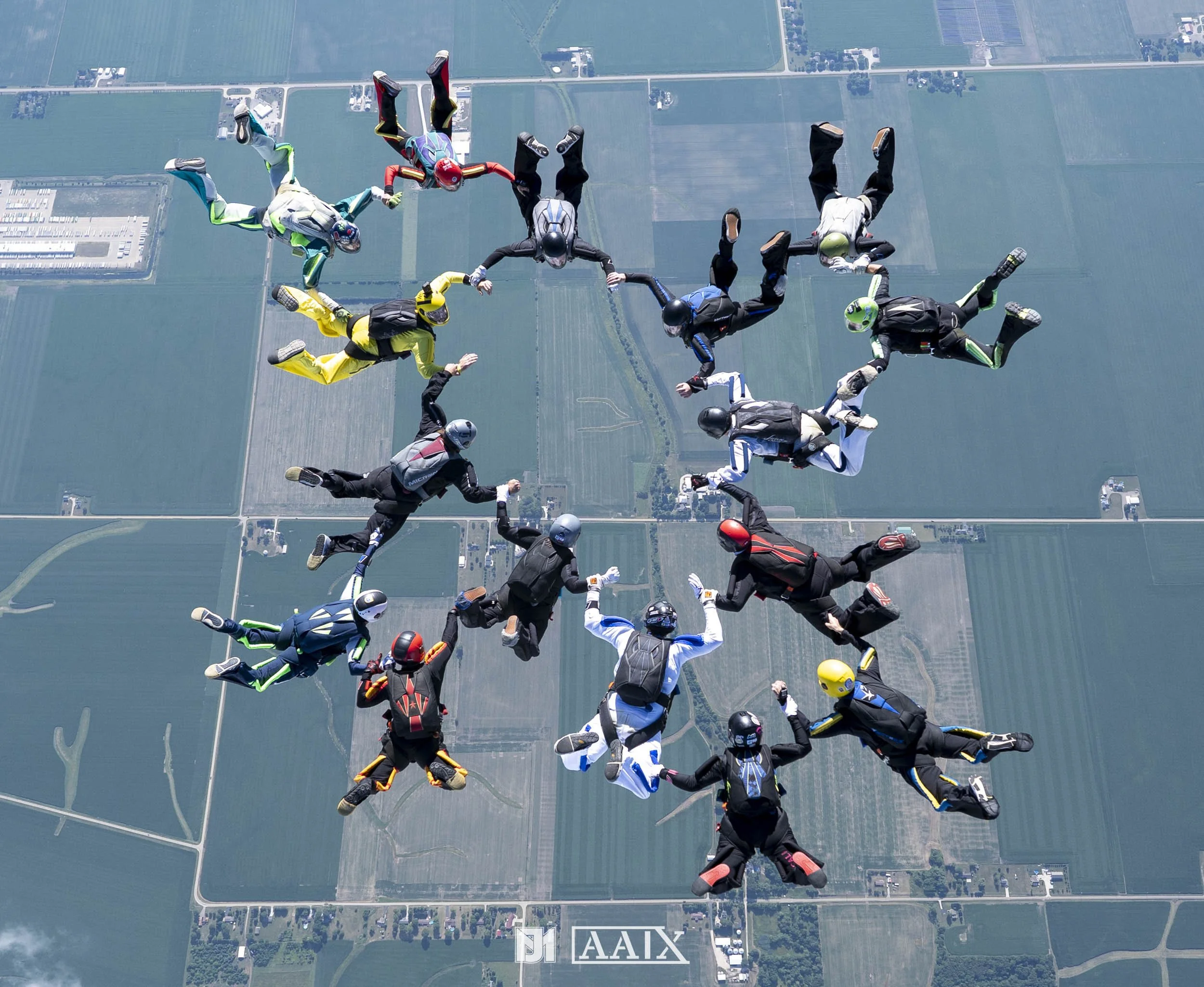 Group of skydivers in colorful jumpsuits and helmets forming a star shape in mid-air above a patchwork of farmland and fields.