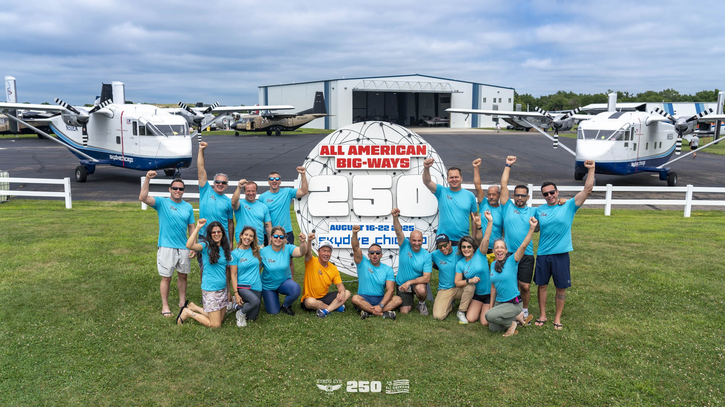 Group of people posing on grass in front of vintage aircrafts and runway, celebrating 250th event of All American Big-Ways skydiving event at Skydive Chicago, August 16-22, 2023.