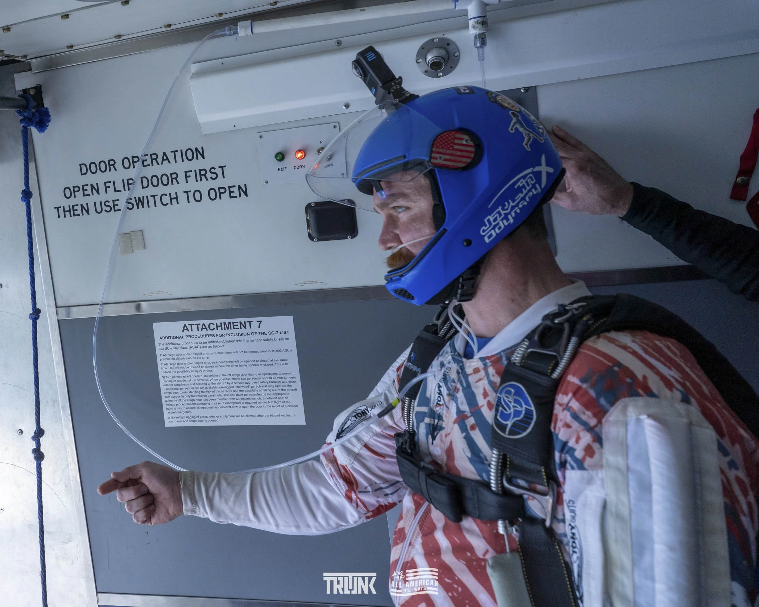 A skydiver wearing a blue helmet, harness, and tiger-striped jumpsuit preparing for a jump. He is inside a plane near the open door, with a sign instructing to open flip door first and use switch to open. A person is helping adjust his helmet.