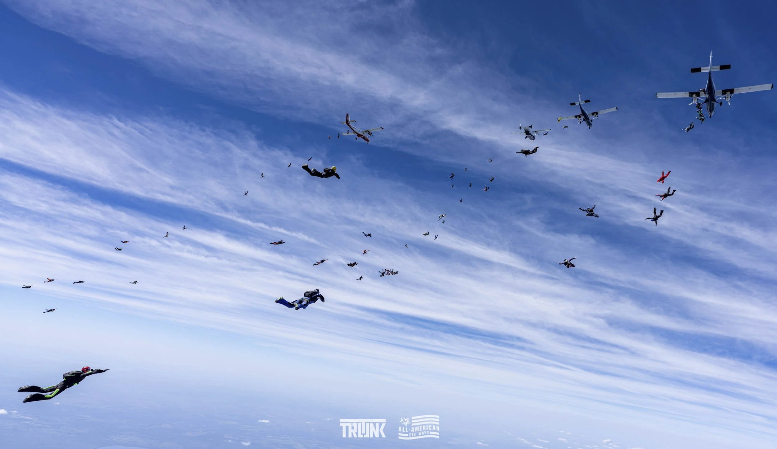 Skydivers in free fall formation with aircraft in the sky and clouds above.