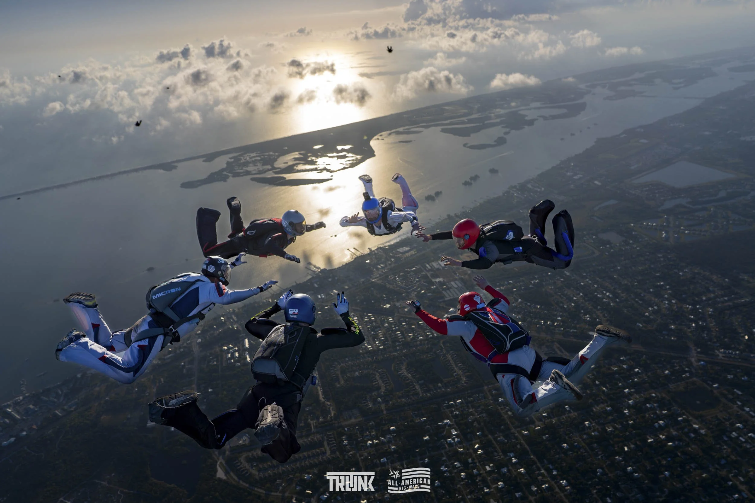 Group of skydivers free-falling in formation over a city and water during sunset.