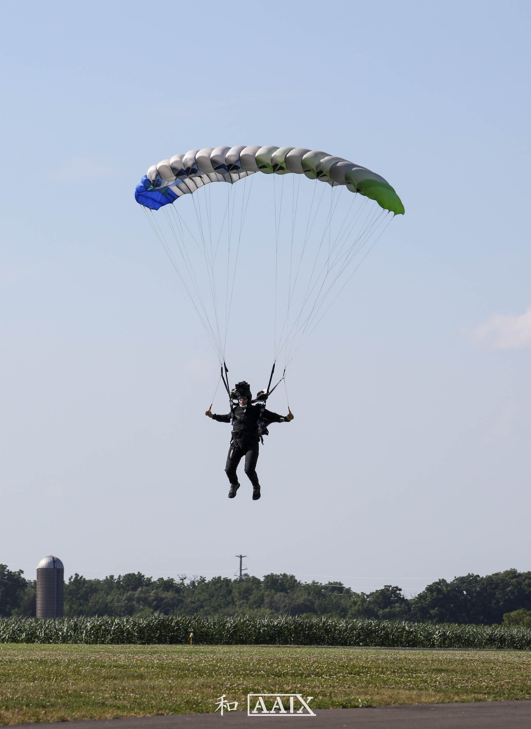 A person parasailing in the sky with a colorful parachute, above a grassy field and trees.