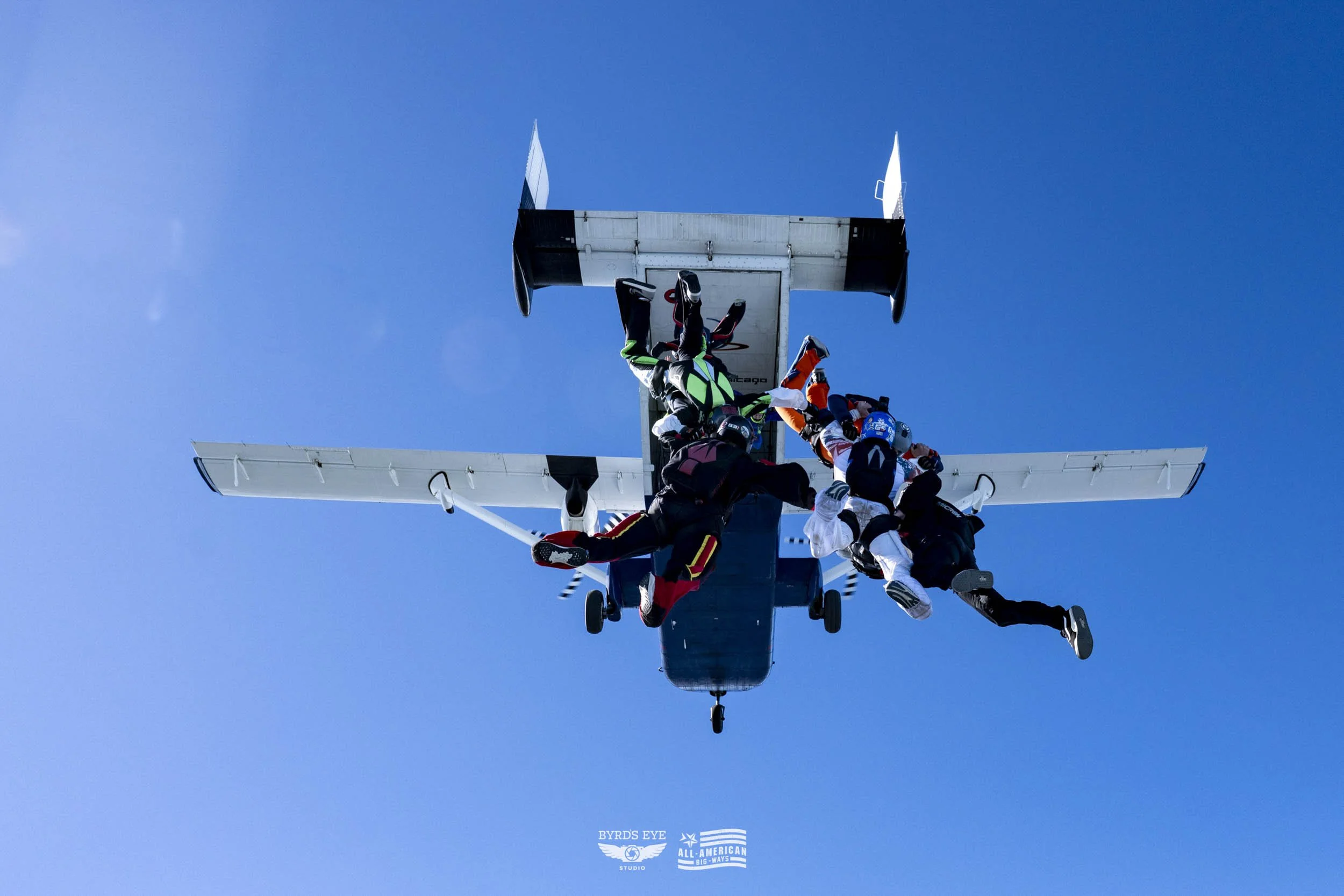 Group of skydivers exiting an aircraft mid-air against a clear blue sky.