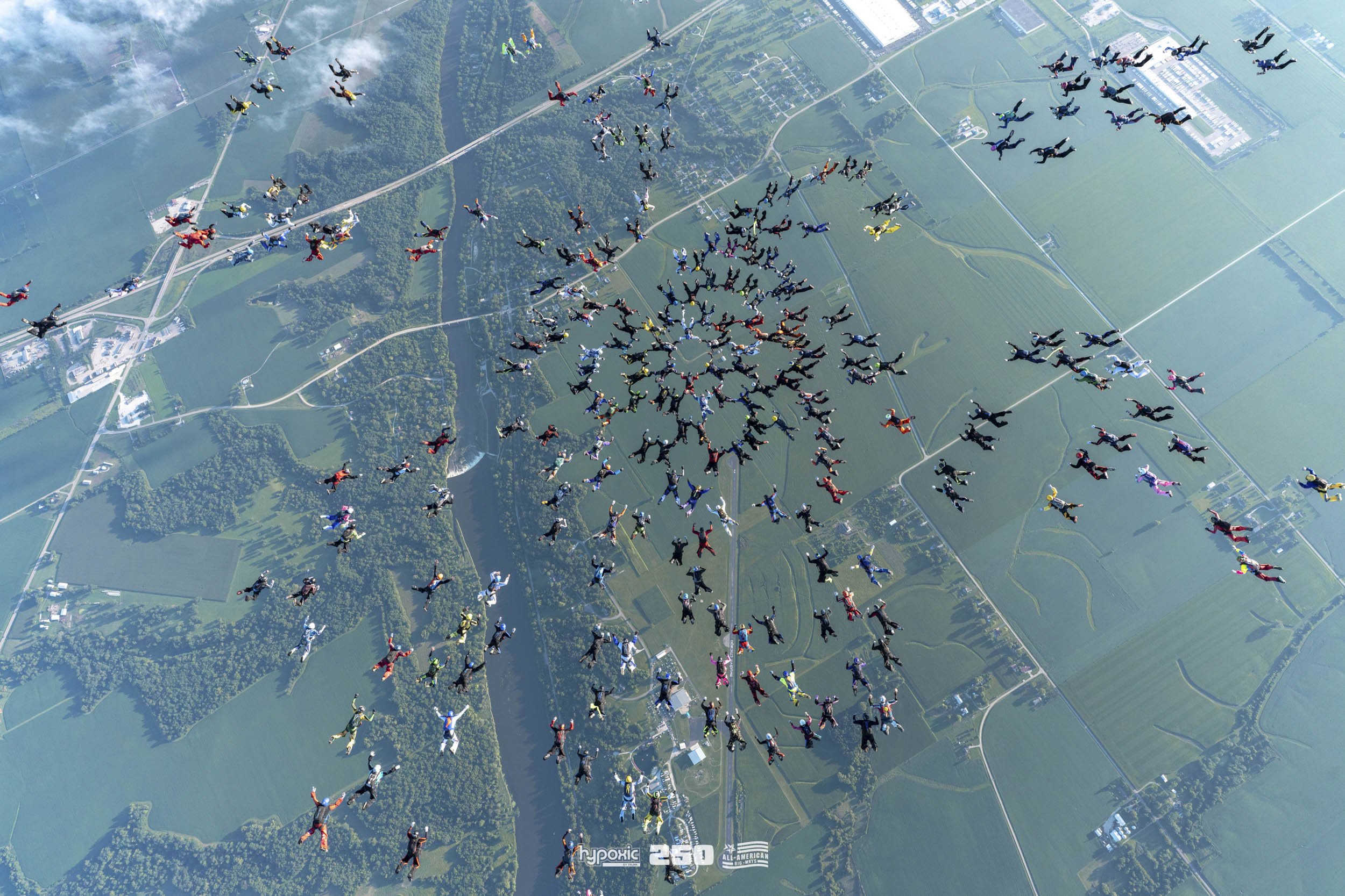 Numerous skydivers in colorful jumpsuits are seen falling in free fall formation over a green landscape with fields and trees.