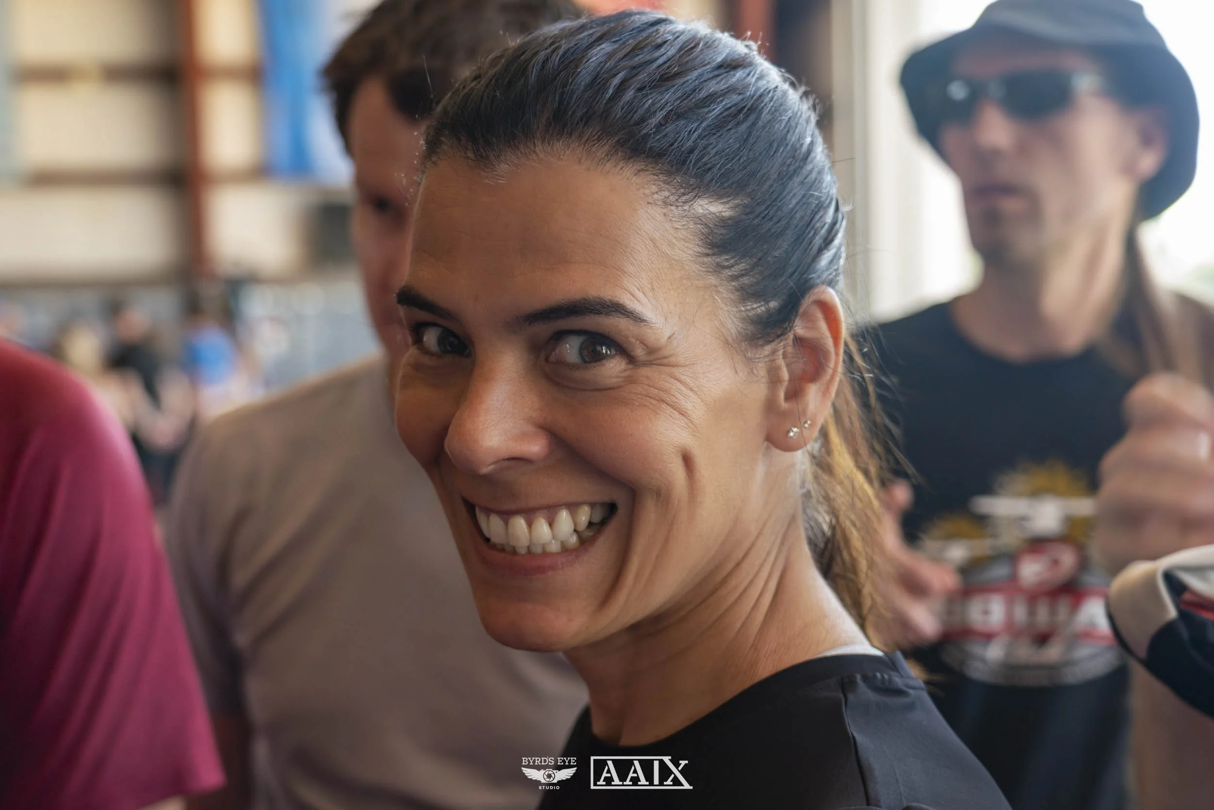 A smiling woman with dark hair pulled back, wearing earrings, in a crowded indoor space with blurred people in the background.