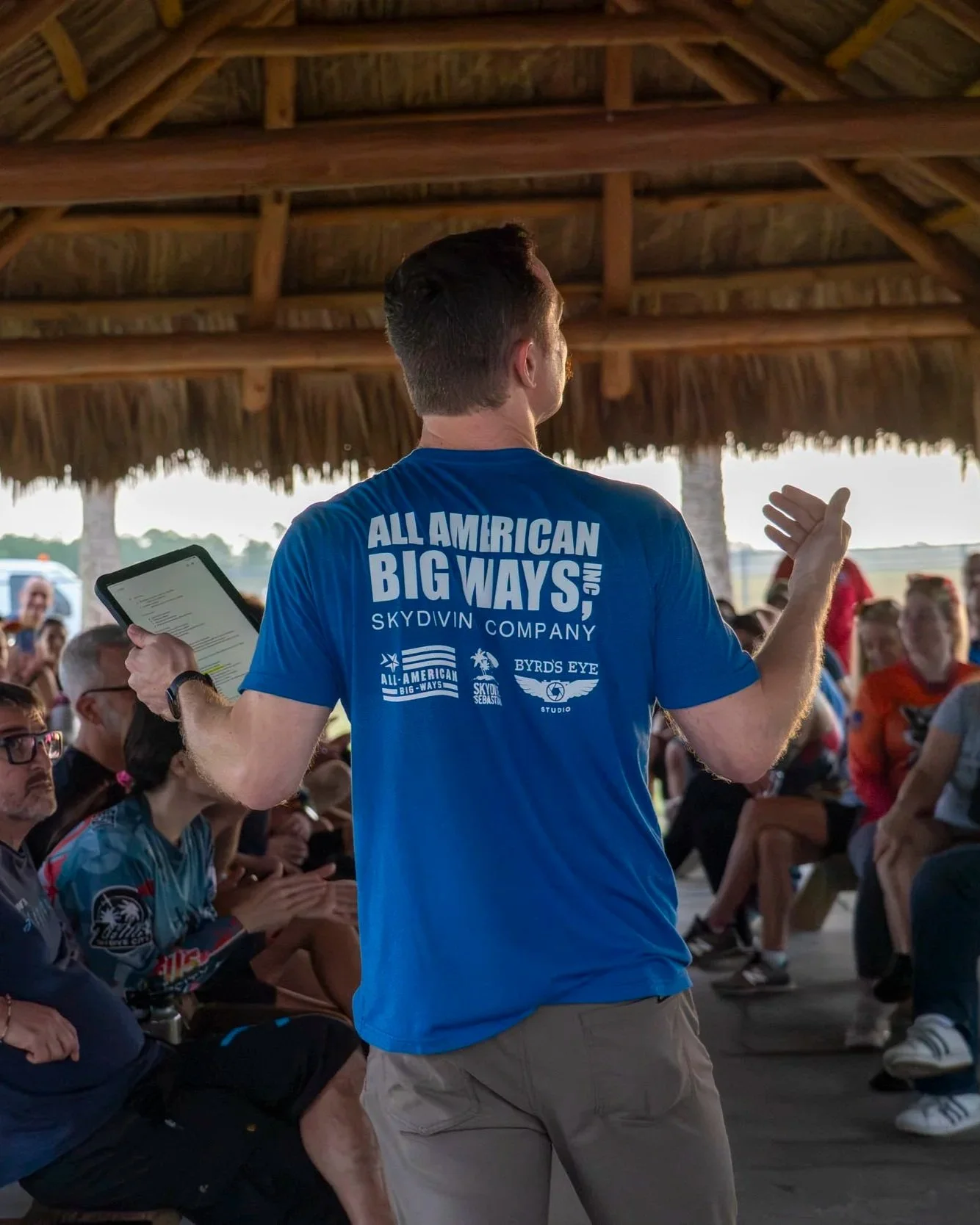 Man wearing a blue t-shirt and khaki shorts giving a presentation or speech to a seated audience in a pavilion with a thatched roof, holding a tablet in his left hand.