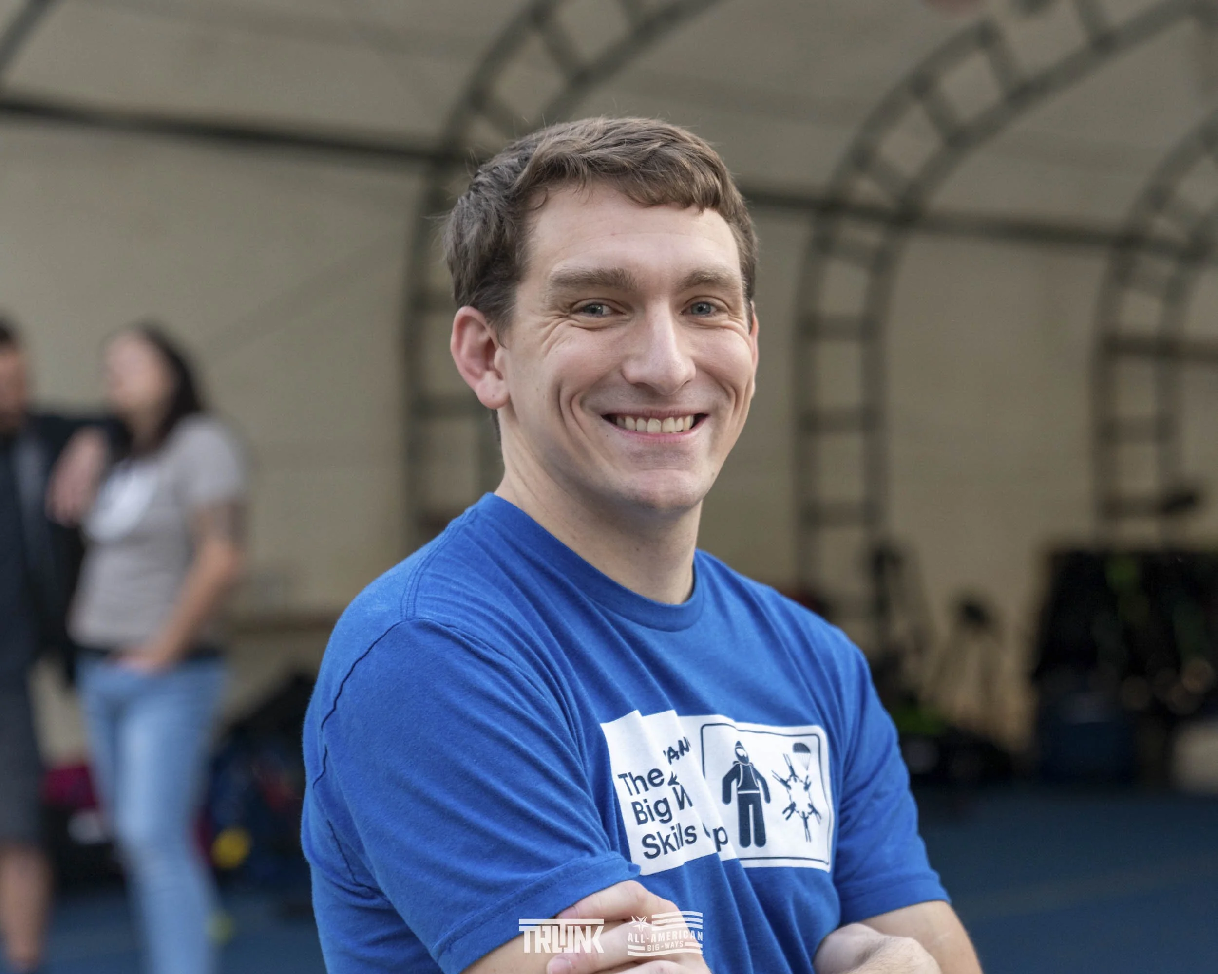 Smiling young man with short brown hair in a blue T-shirt with text and graphics, standing indoors with blurred people in the background.
