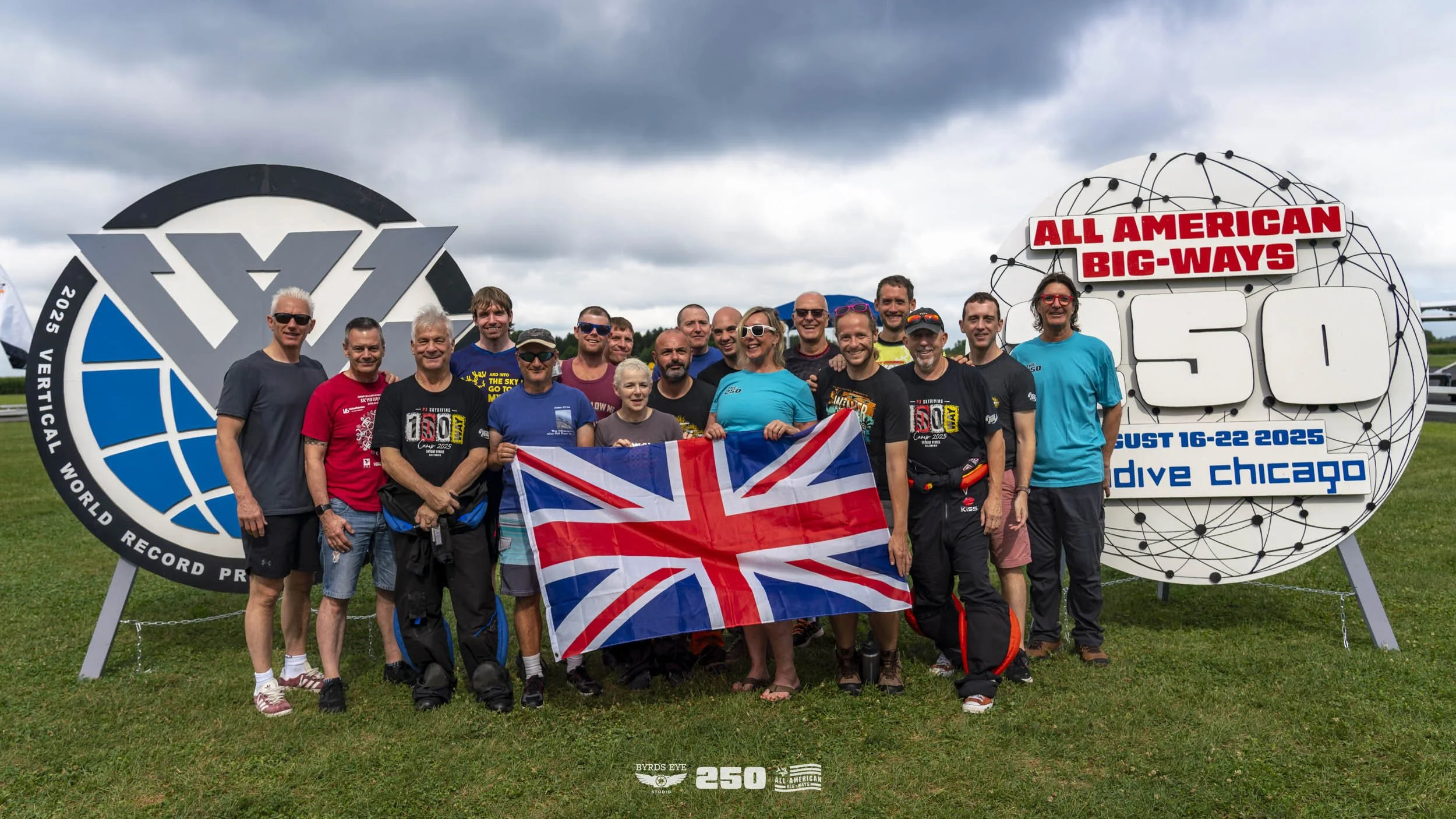 Group of 17 people standing outdoors on grass, holding a UK flag, posing for a photo in front of sculptures and signs celebrating the 250th anniversary of the all-American Big-Ways Skydiving event in Chicago, August 16-22, 2025.