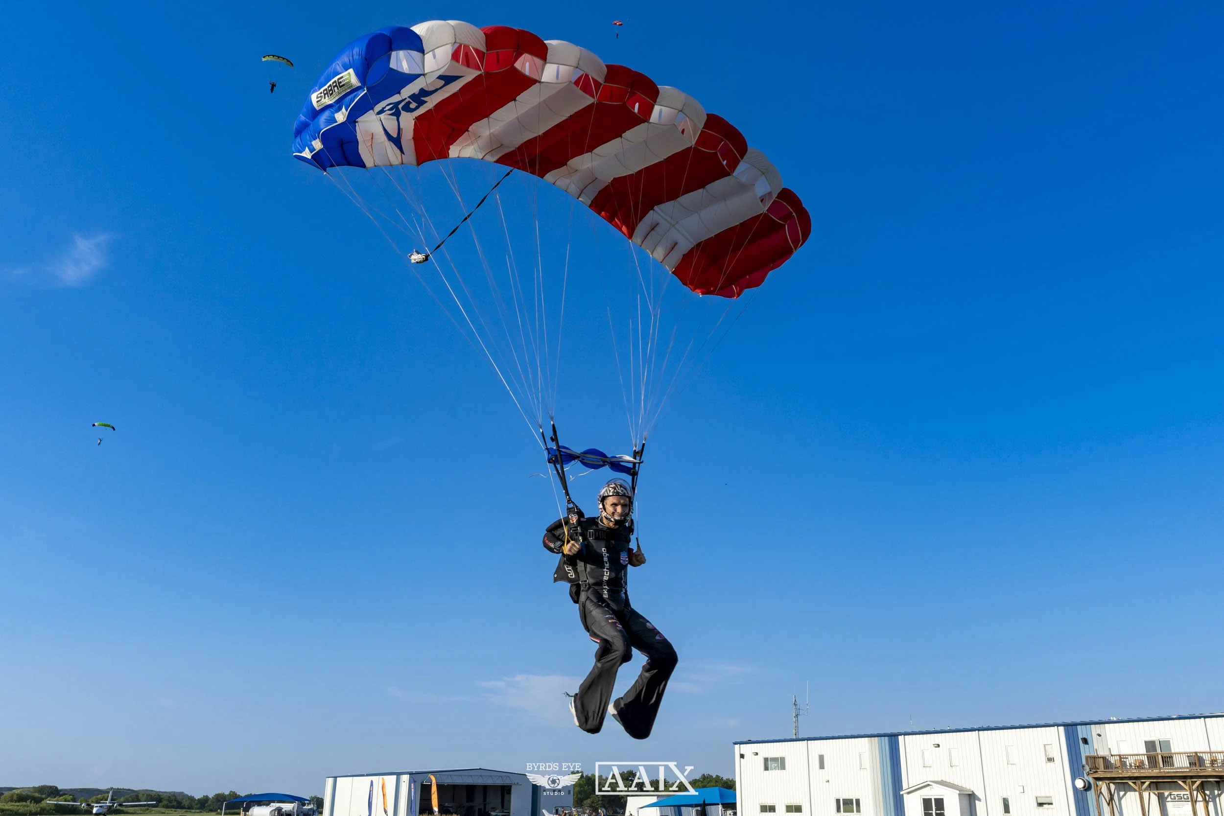 A person paragliding with a red, white, and blue canopy in a clear blue sky, with other paragliders visible in the distance.
