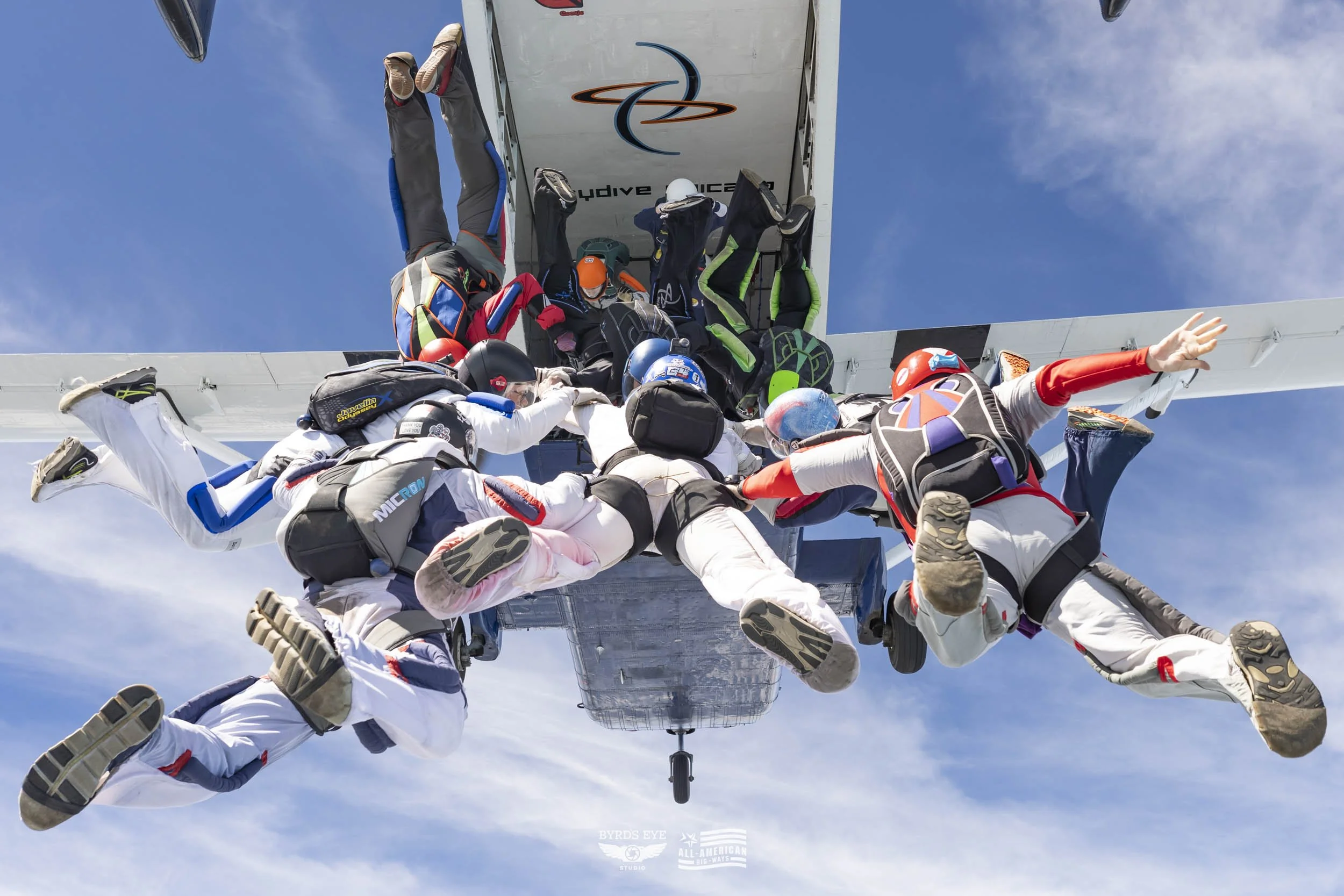 Skydivers holding onto a descending airplane, preparing to jump from the plane in mid-air against a blue sky.