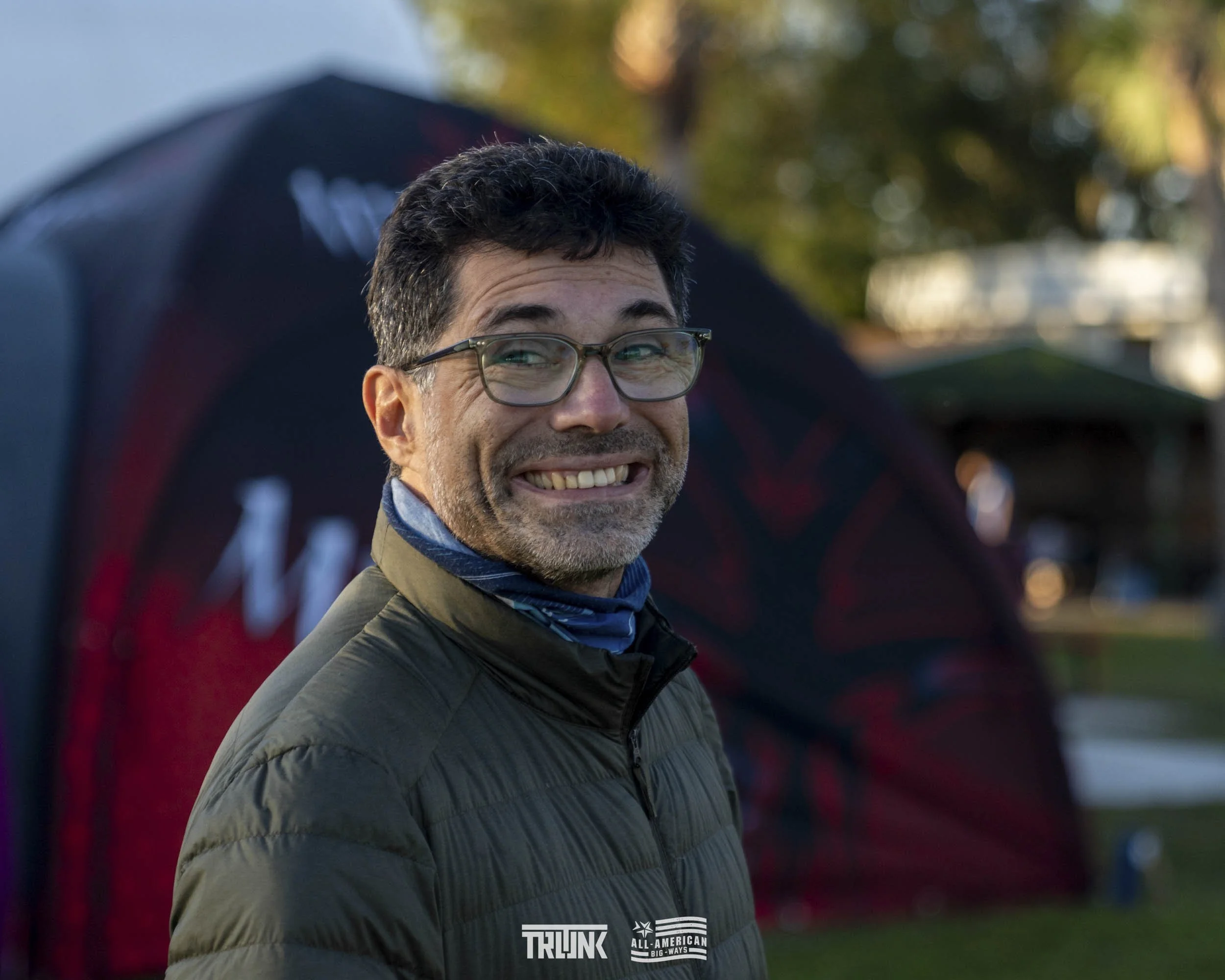 A man with glasses and dark curly hair smiling outdoors, wearing a dark quilted jacket with a blue neck gaiter, standing in front of a blurred red and black tent with trees and a park-like setting in the background.