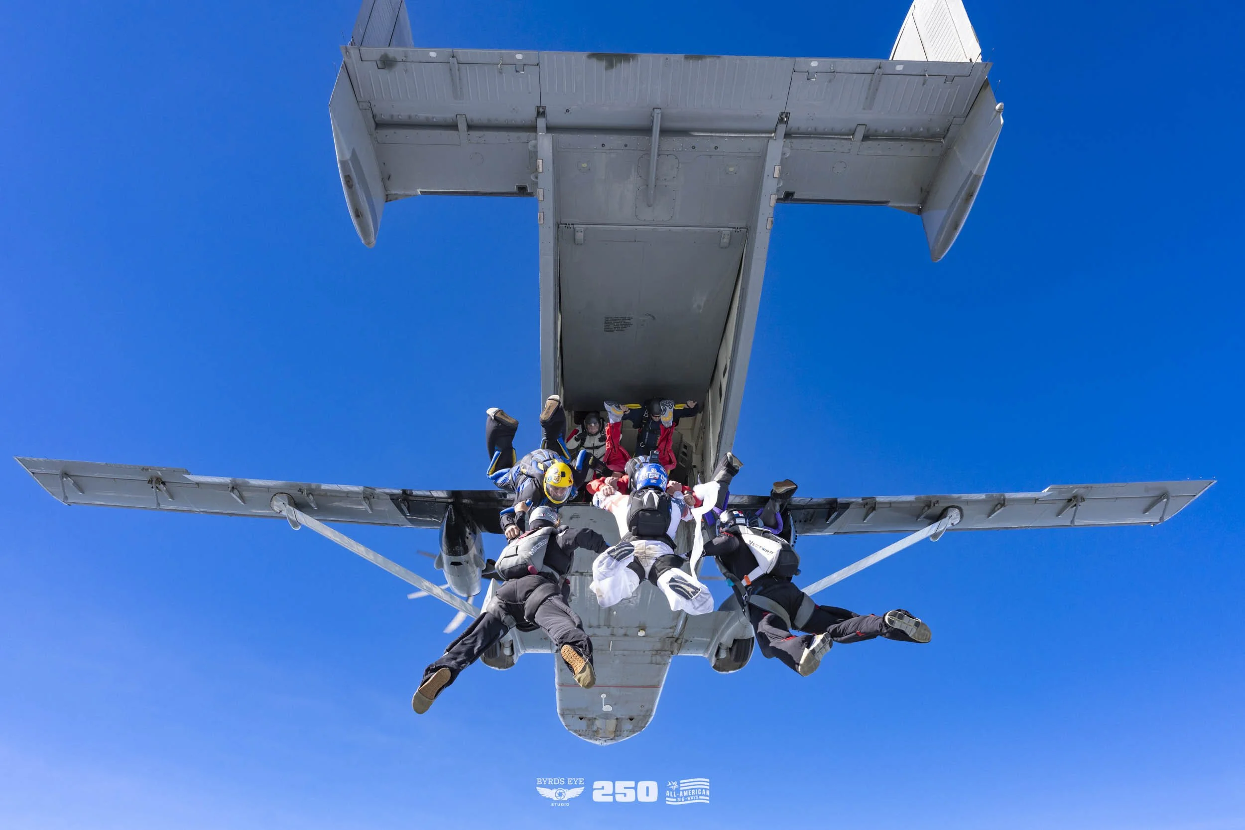Seven skydivers exit an airplane with blue sky in the background, holding on to the airplane's open door during freefall.