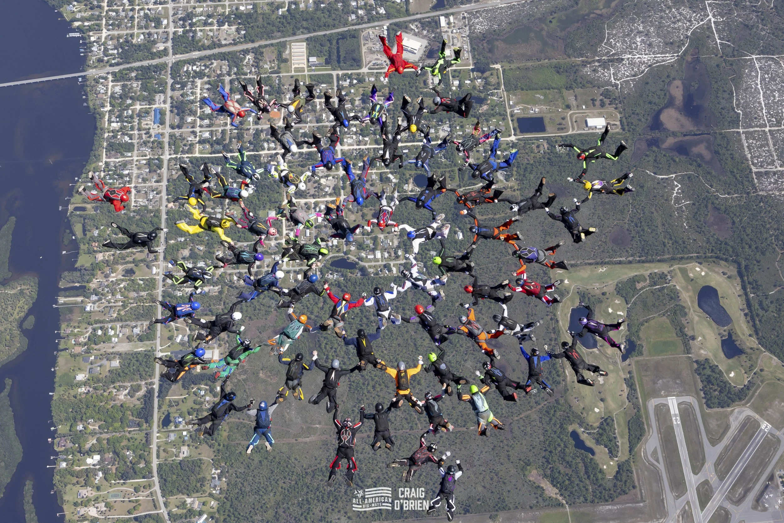 A large group of skydivers in colorful jumpsuits and helmets are skydiving in a formation over a landscape that includes a river, roads, and parks.