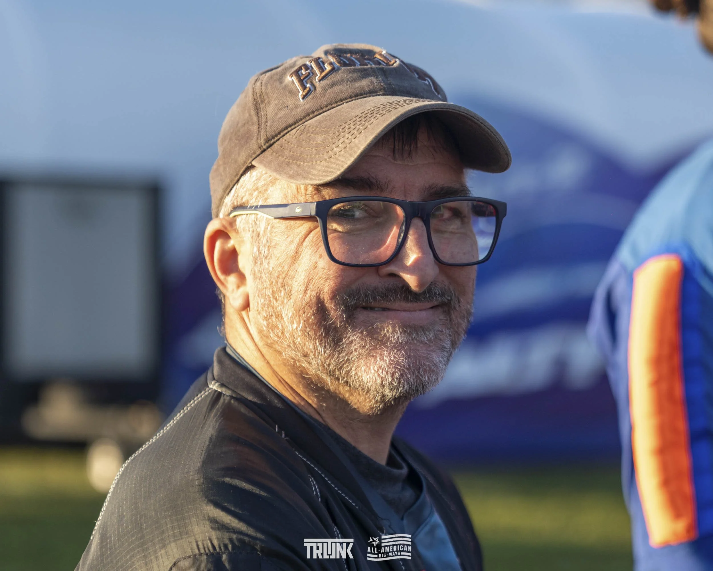 A middle-aged man with glasses and a gray beard smiling outdoors, wearing a baseball cap and black jacket, with a blue tent and people in the background.