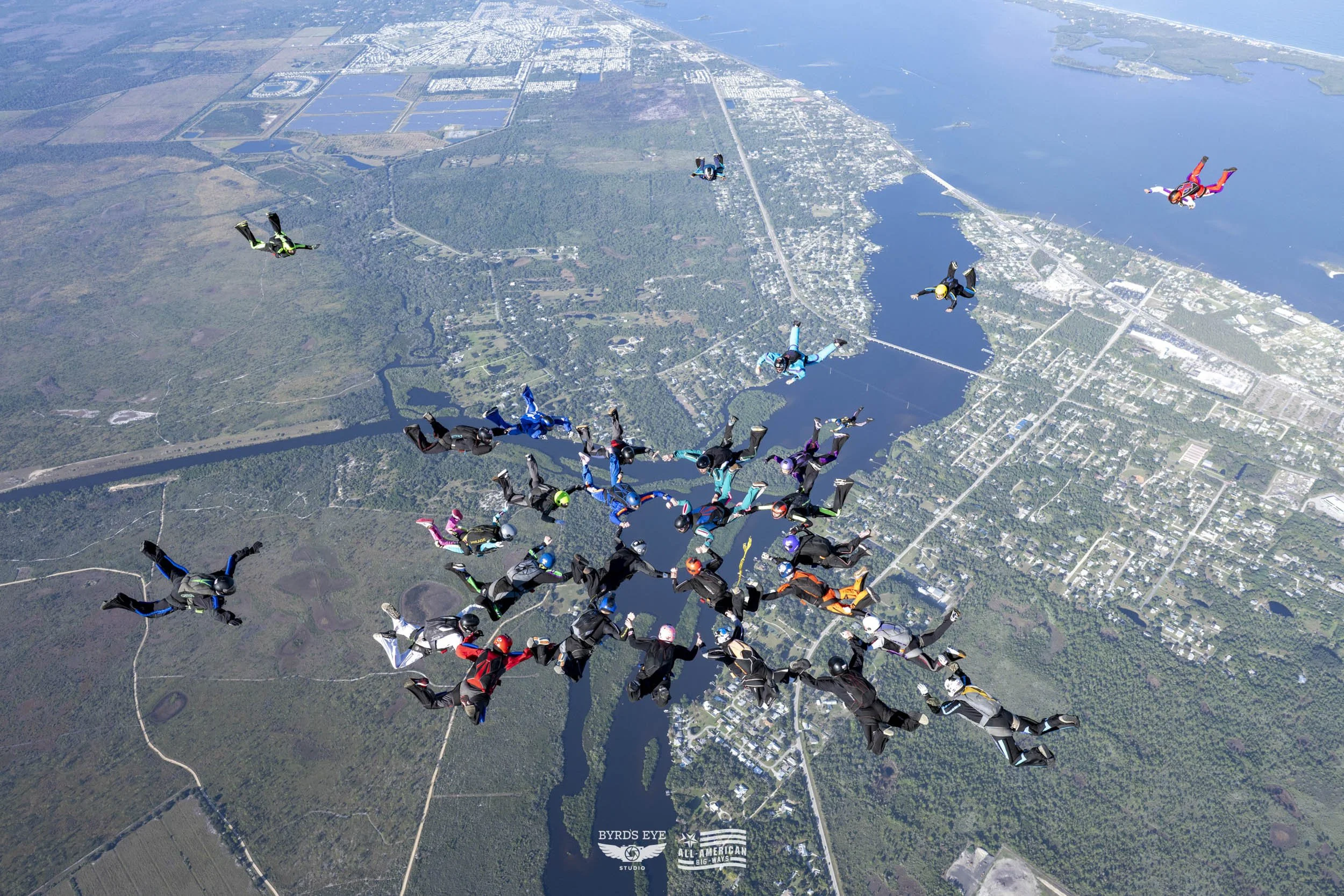 Group of skydivers in free fall over a landscape with lakes, forests, and a city in the background.