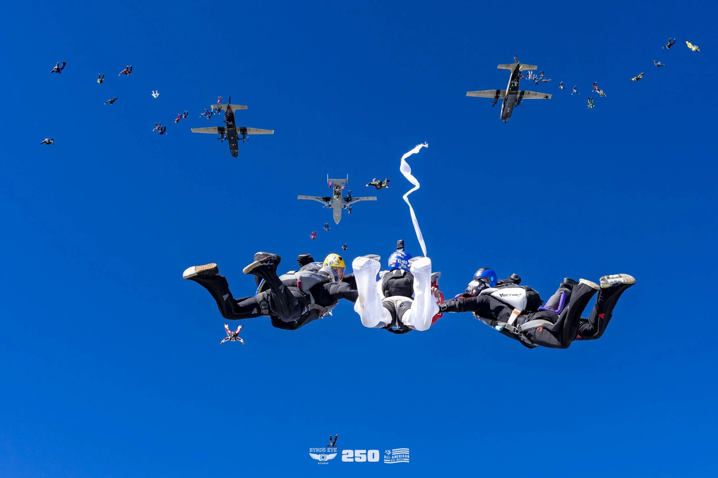 Skydivers in free fall formation with aircraft flying overhead against a clear blue sky.