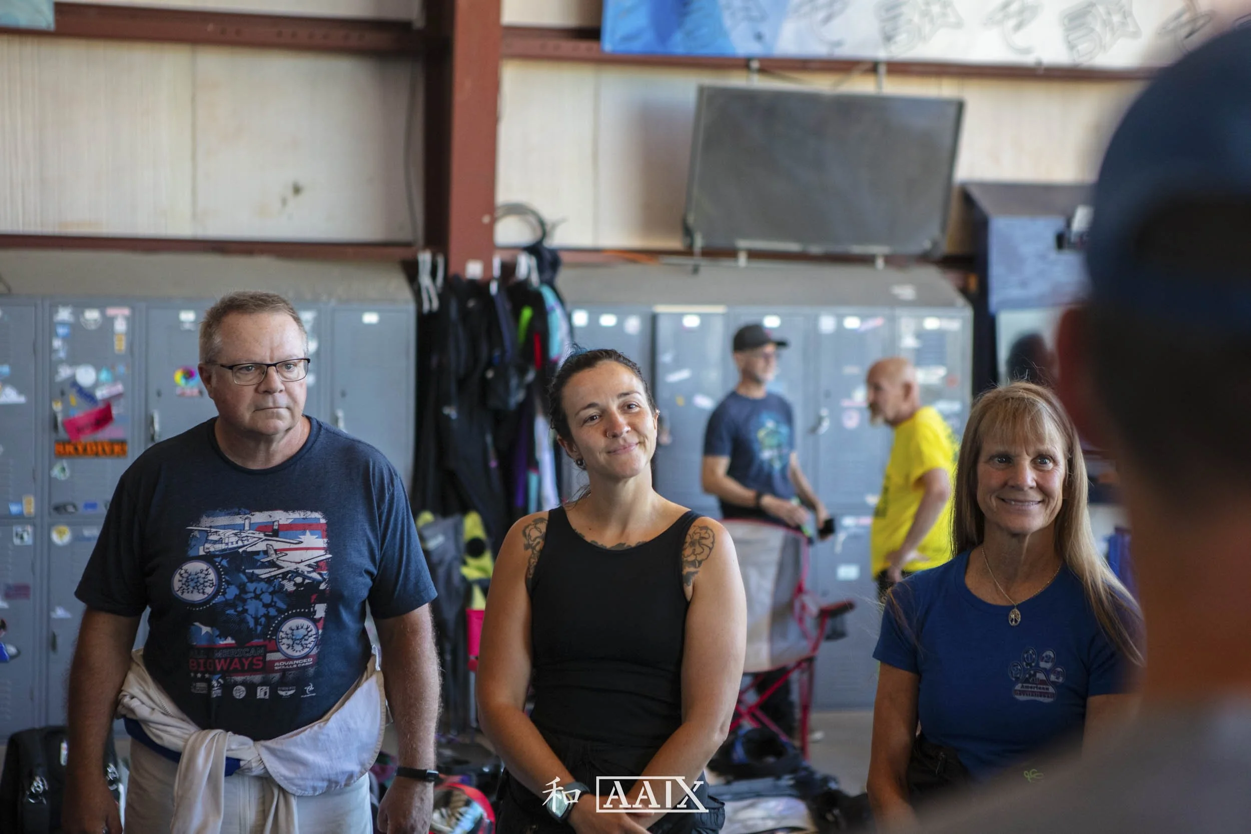 Group of four people standing in a locker room, with lockers and backpacks behind them, and two people in the background engaged in conversation.