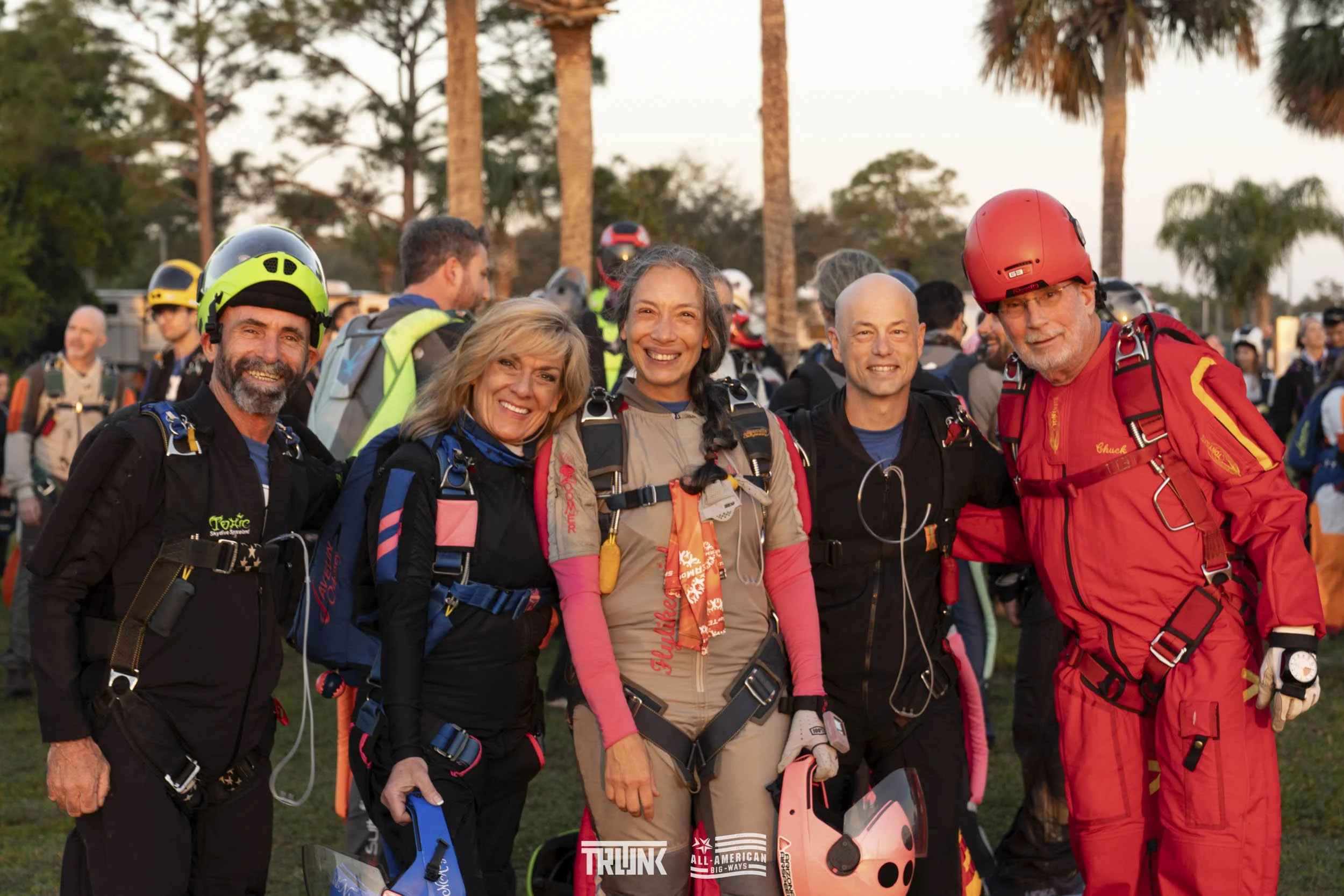 Group of five smiling skydivers in jumpsuits and helmets, standing outdoors with palm trees and other skydivers in the background during sunset.
