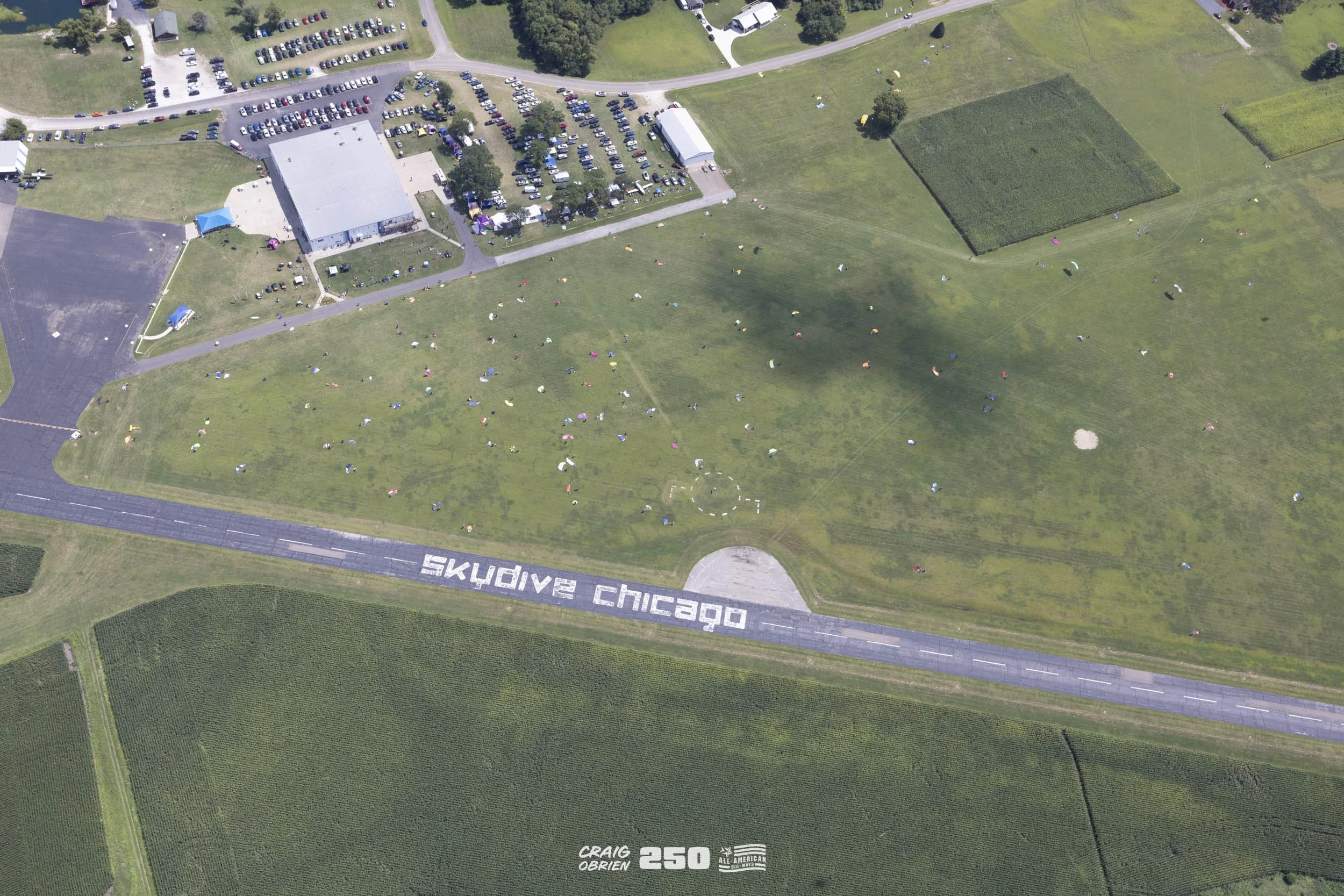 Overhead view of a park with small groups of people scattered across the grassy area, some participating in an event. A large road with the words 'SKYDIVE CHICAGO' painted on it runs horizontally across the bottom of the image. There are several buil