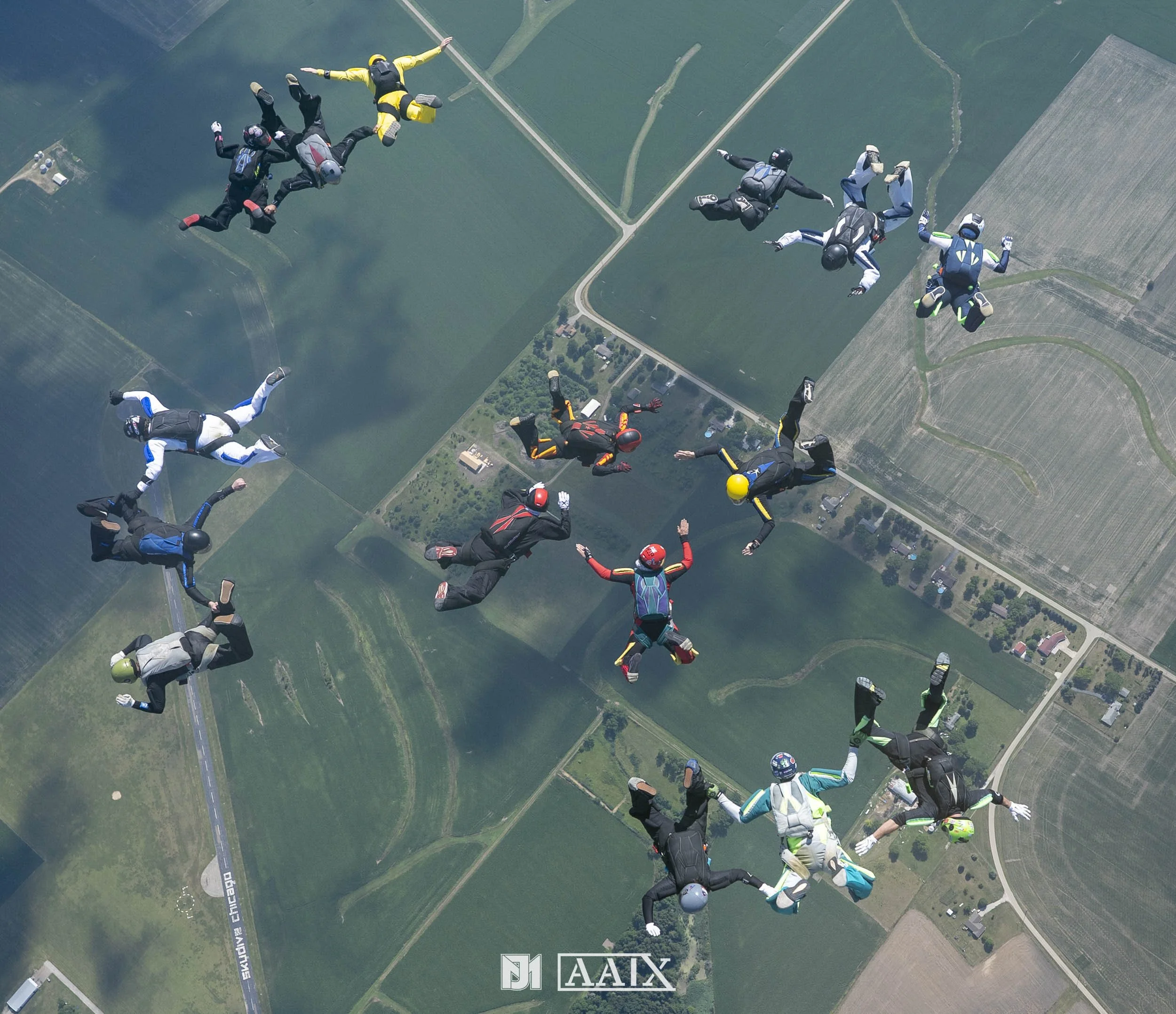 Group of skydivers in free fall over a rural landscape with fields and roads.