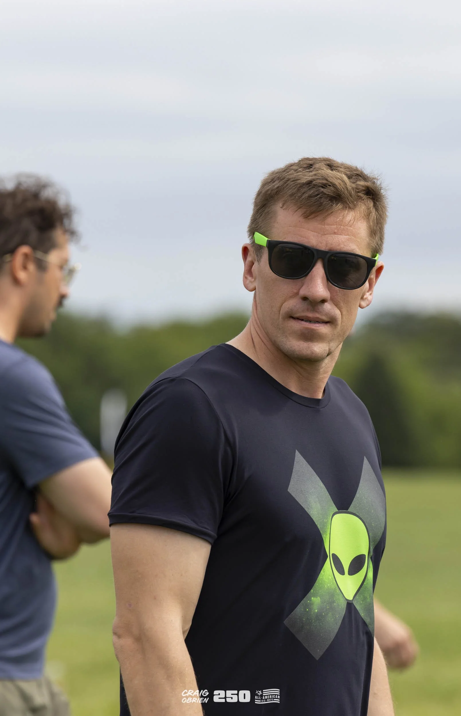 A man with short red hair wearing black sunglasses with green accents and a black T-shirt with a green alien head logo stands outdoors on a cloudy day, with blurred greenery and another person in the background.