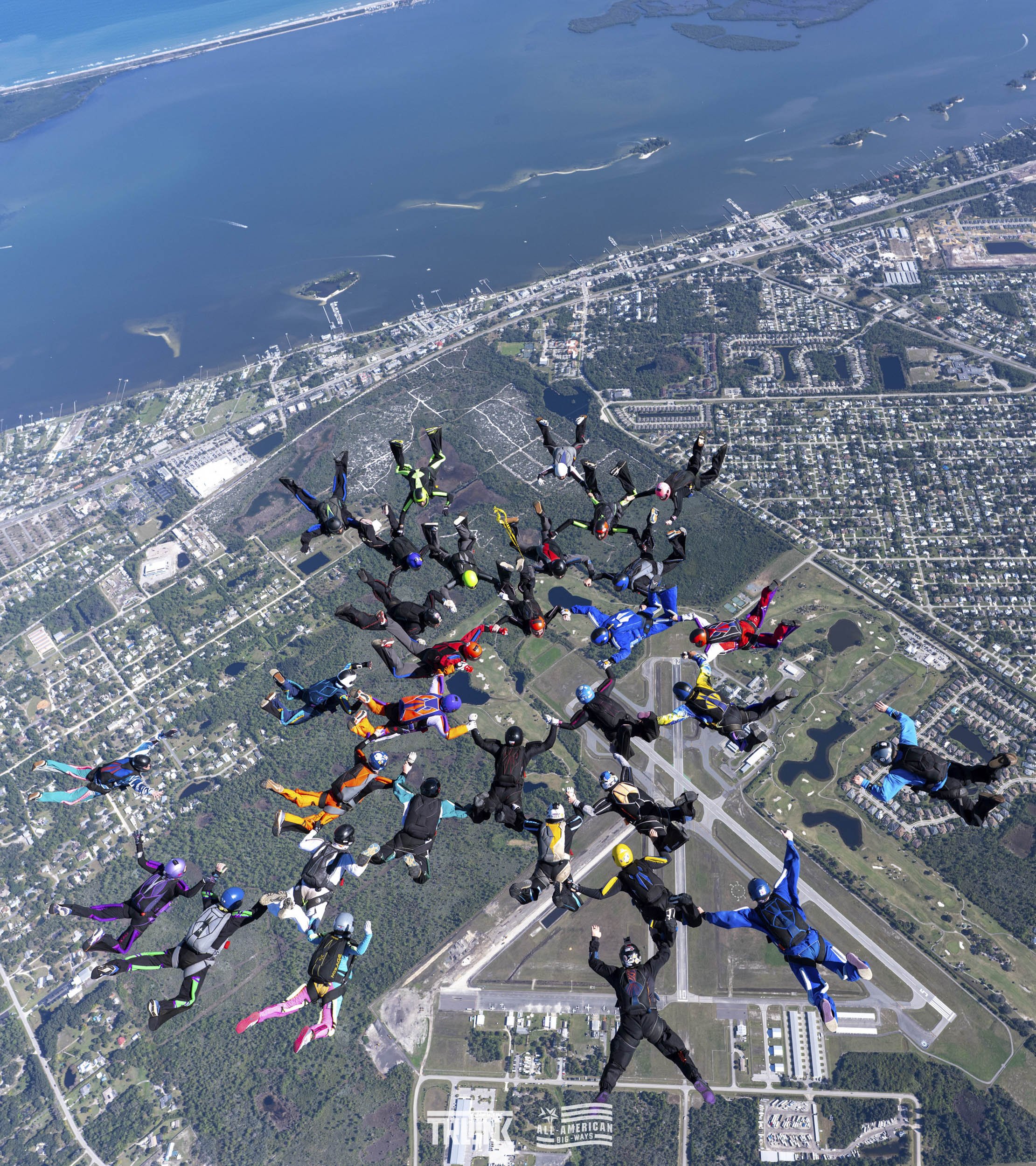 A group of skydivers in colorful jumpsuits and helmets floating in a formation above a city with water and roads visible below.