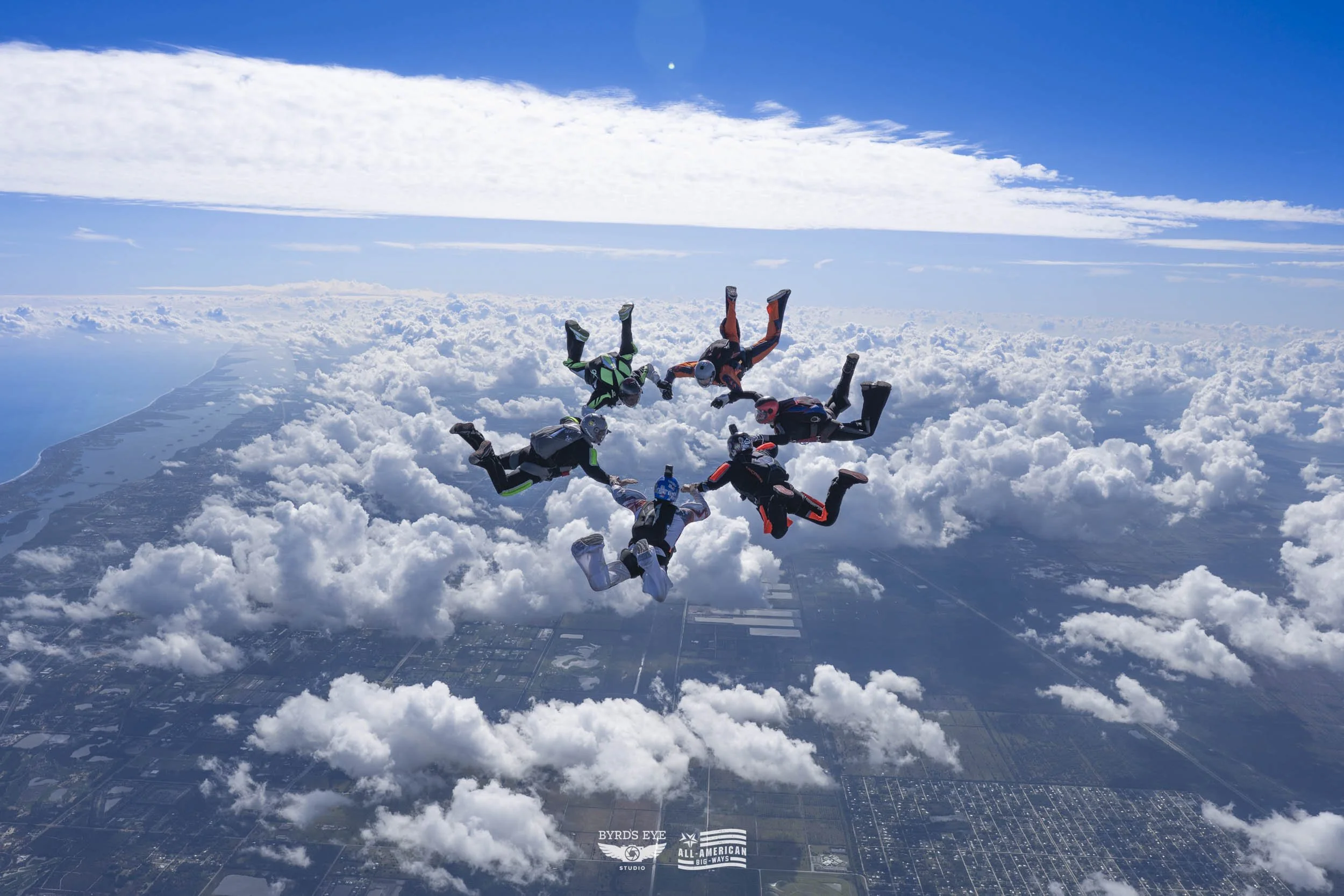 Six skydivers in jumpsuits and helmets forming a circle in a free-fall formation above clouds and a landscape, with a bright blue sky and a moon visible in the background.