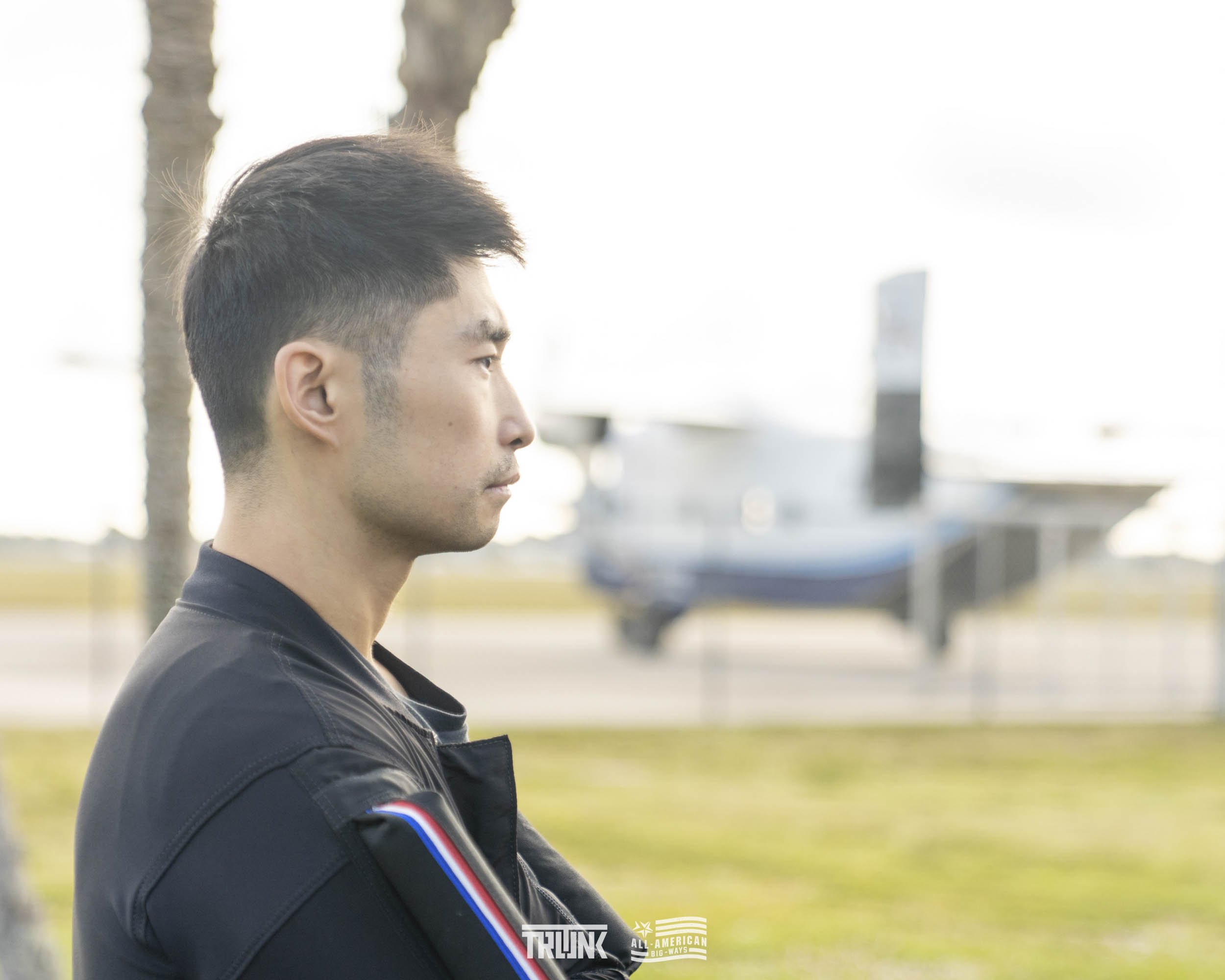 A man in profile, wearing a black jacket, stands outdoors with trees and a boat in the background.