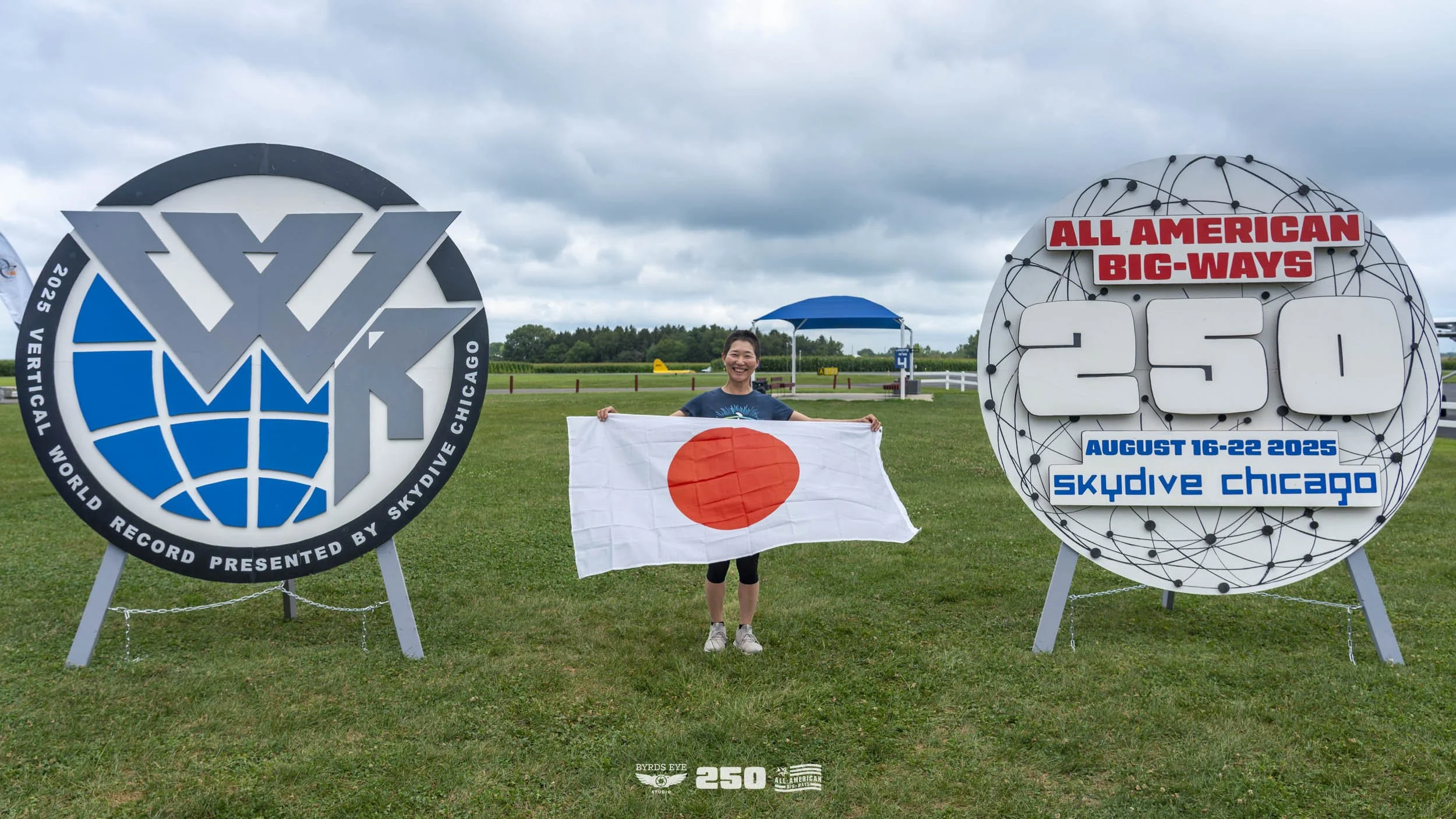 A woman smiling and holding a Japanese flag stands between two large signs at Skydive Chicago. The left sign features the 2025 World Record logo, and the right sign displays information about the All American Big-ways event, including the dates Augus