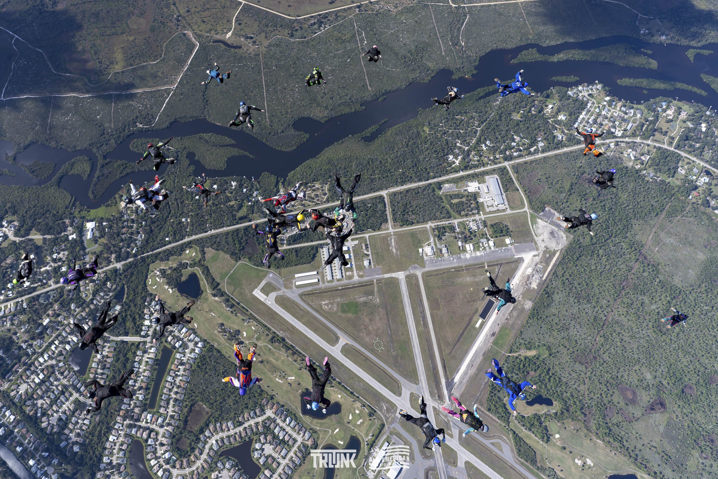 Skydivers in free fall above a landscape with lakes, forests, roads, and buildings.