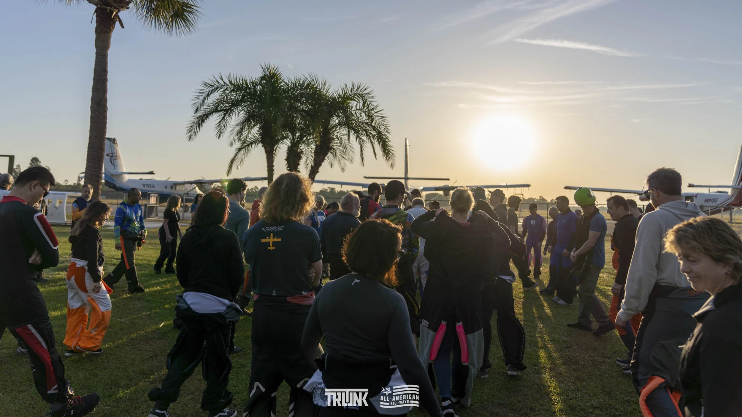 A crowd of people gathered on a grassy field near small airplanes at sunset, with palm trees and a clear sky in the background.