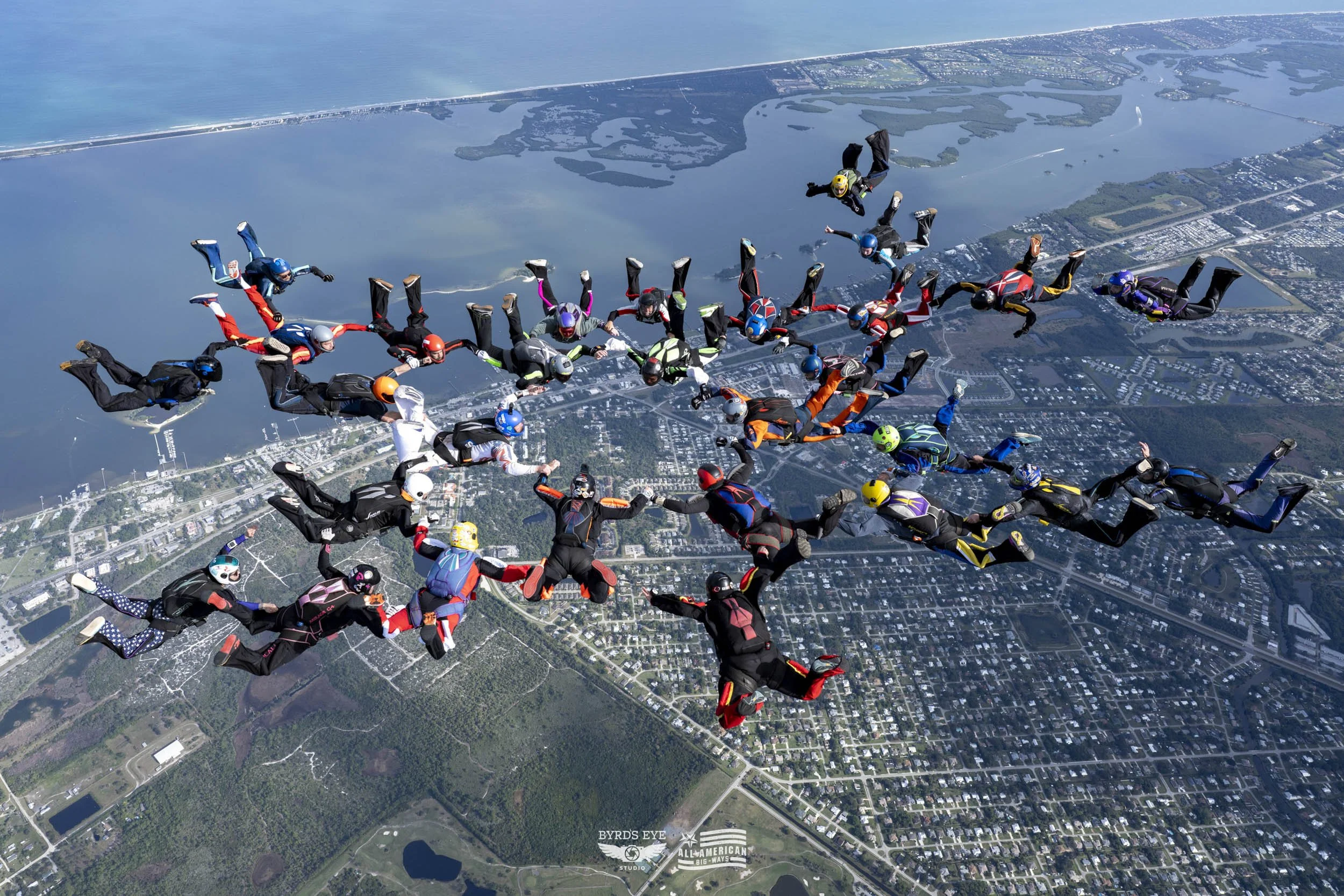 Group of skydivers in freefall formation over a landscape with water, roads, and forests.