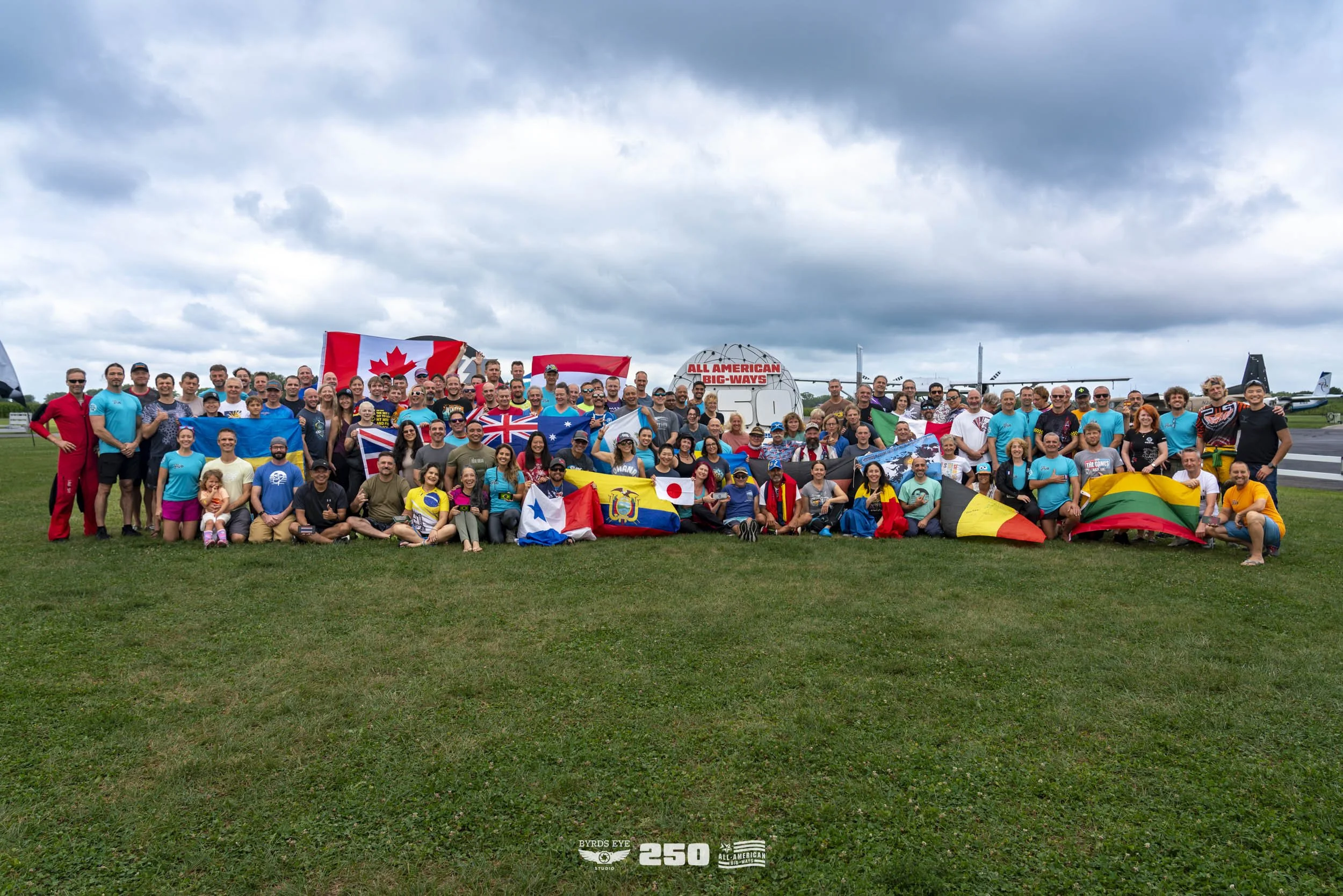 Large group of people gathered on grass at an airfield, holding various flags from different countries, with airplanes in the background under cloudy skies.