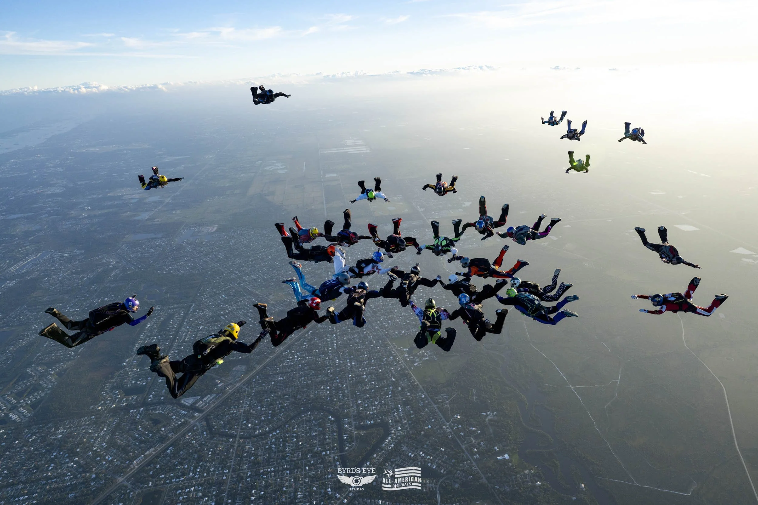 Group of skydivers in freefall formation during a jump, with a city and landscape visible below.