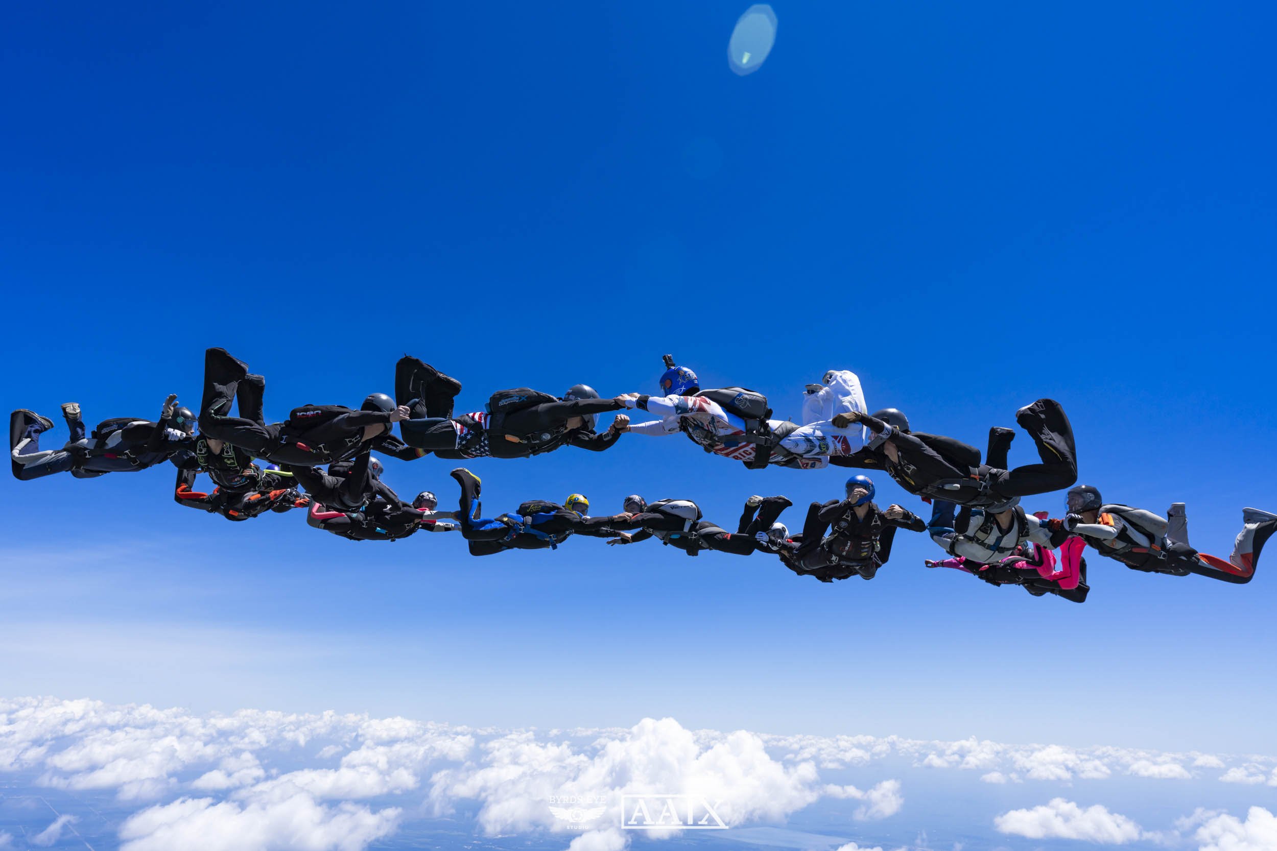 A group of skydivers holding hands in a formation, flying high above the clouds against a clear blue sky.