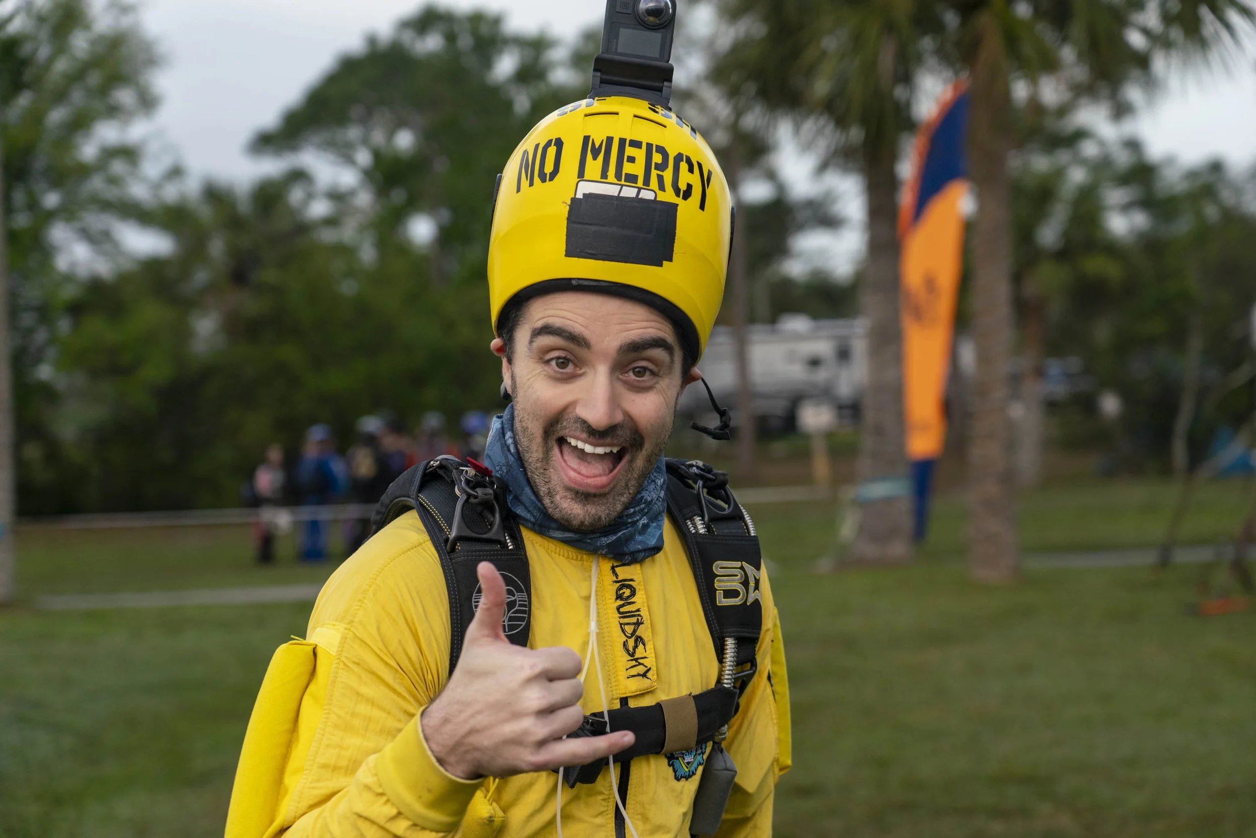 A man smiling and giving a thumbs-up while wearing a yellow helmet with the words 'NO MERCY' written on it, and a yellow jacket, outdoors with trees and flags in the background.